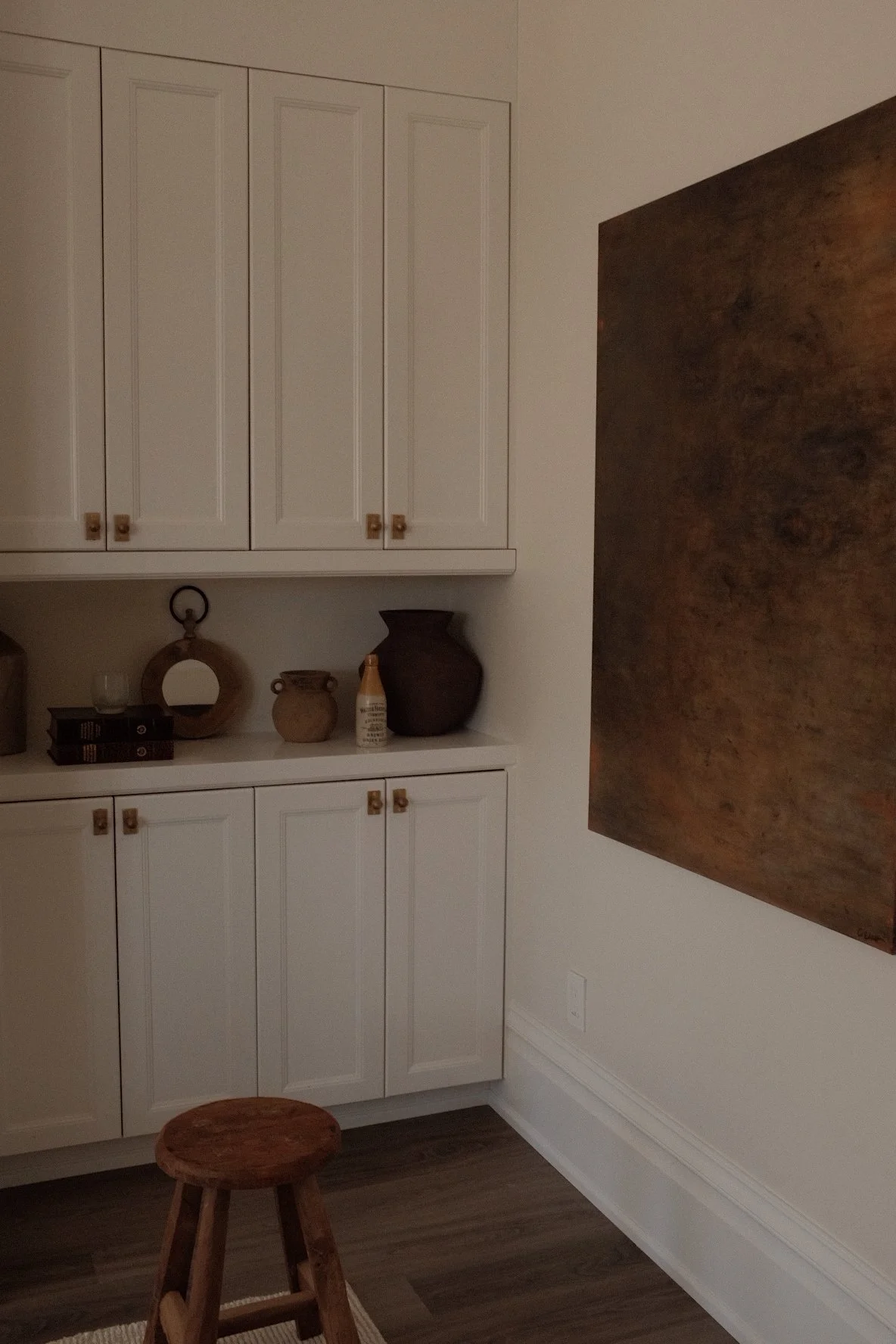 A corner of a room with white cabinets, decorative pottery, and a large abstract painting on the wall. A small wooden stool is on a rug on dark wood flooring.