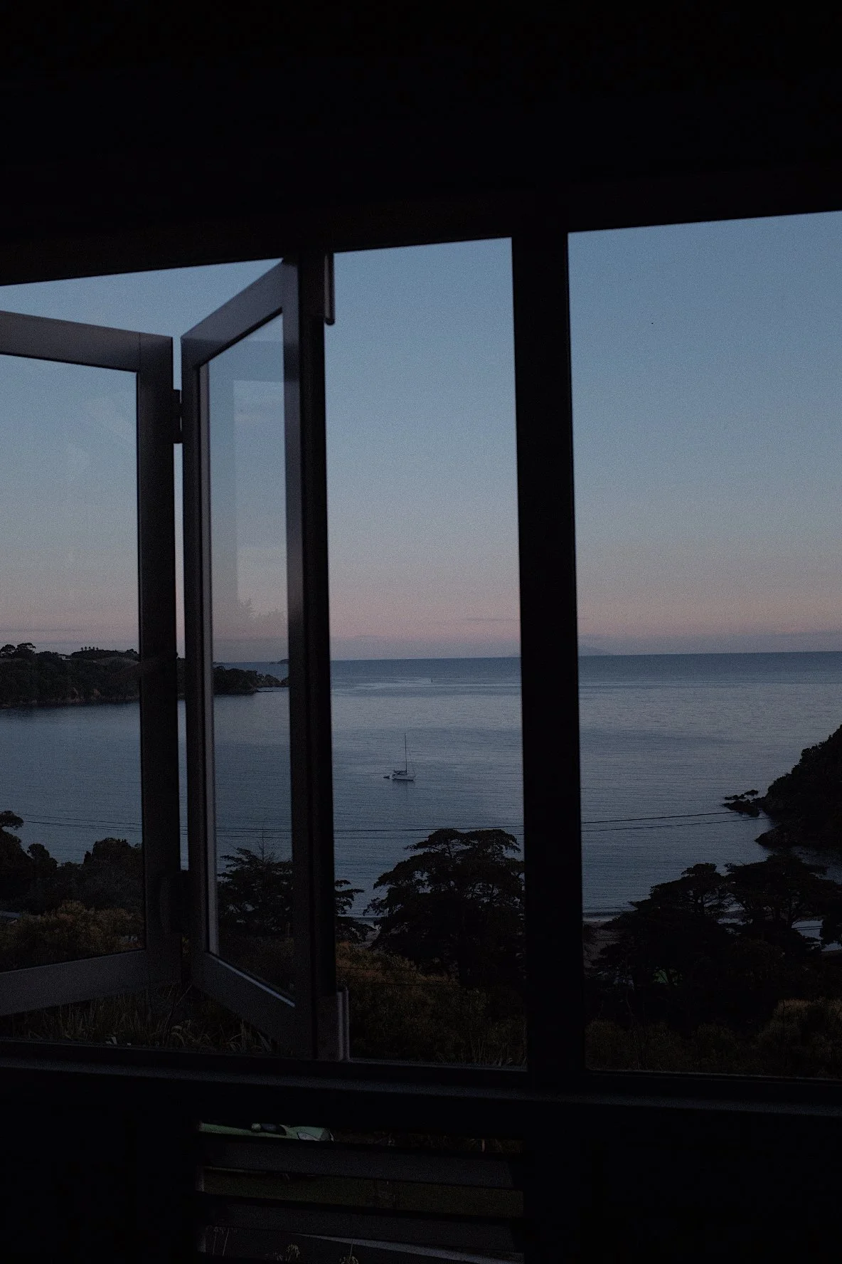 Open window with ocean view during sunset, sailboat on water, and silhouette of trees.