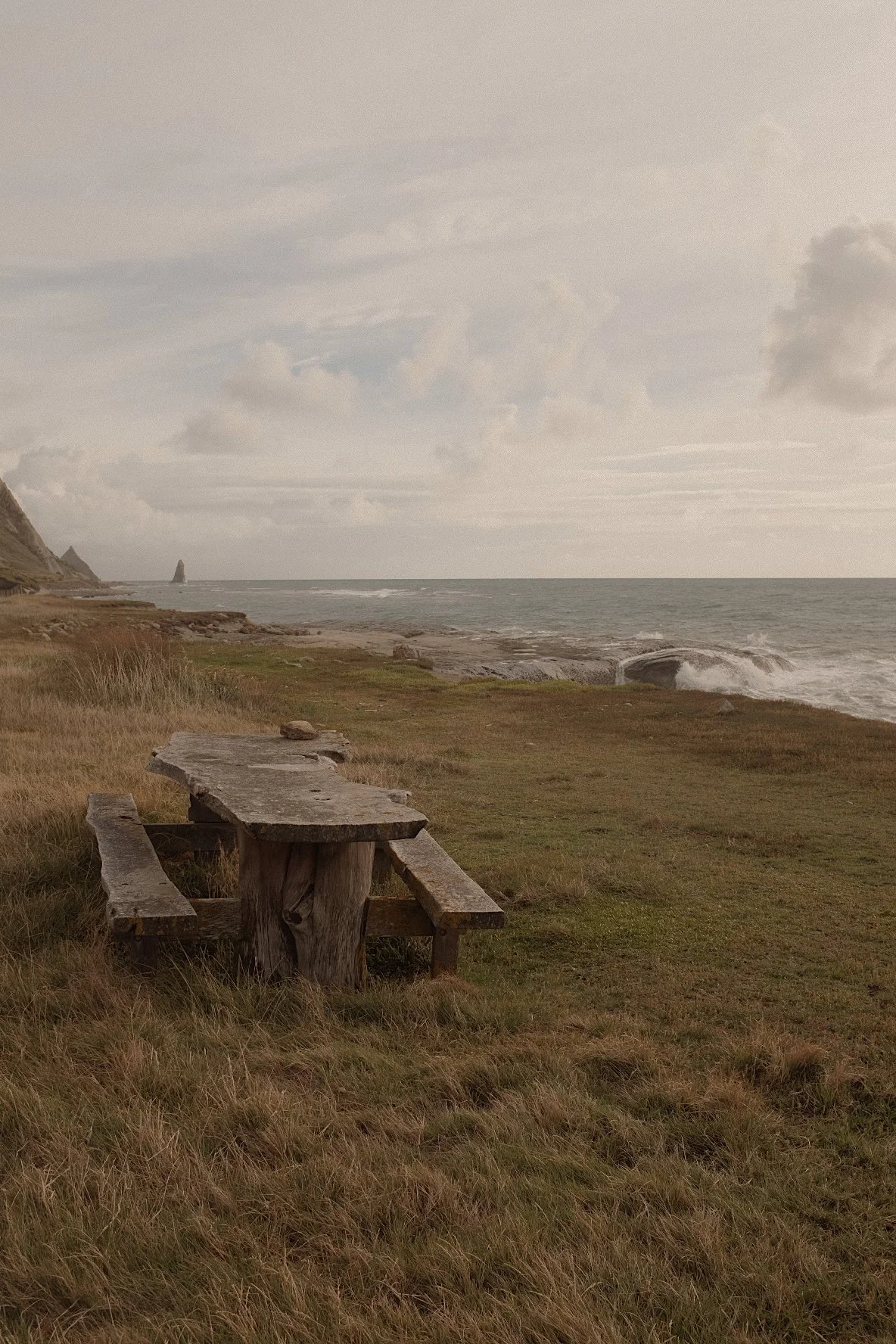 Rustic wooden picnic bench on grassy coastal landscape, with the ocean and cliffs in the background under a cloudy sky.