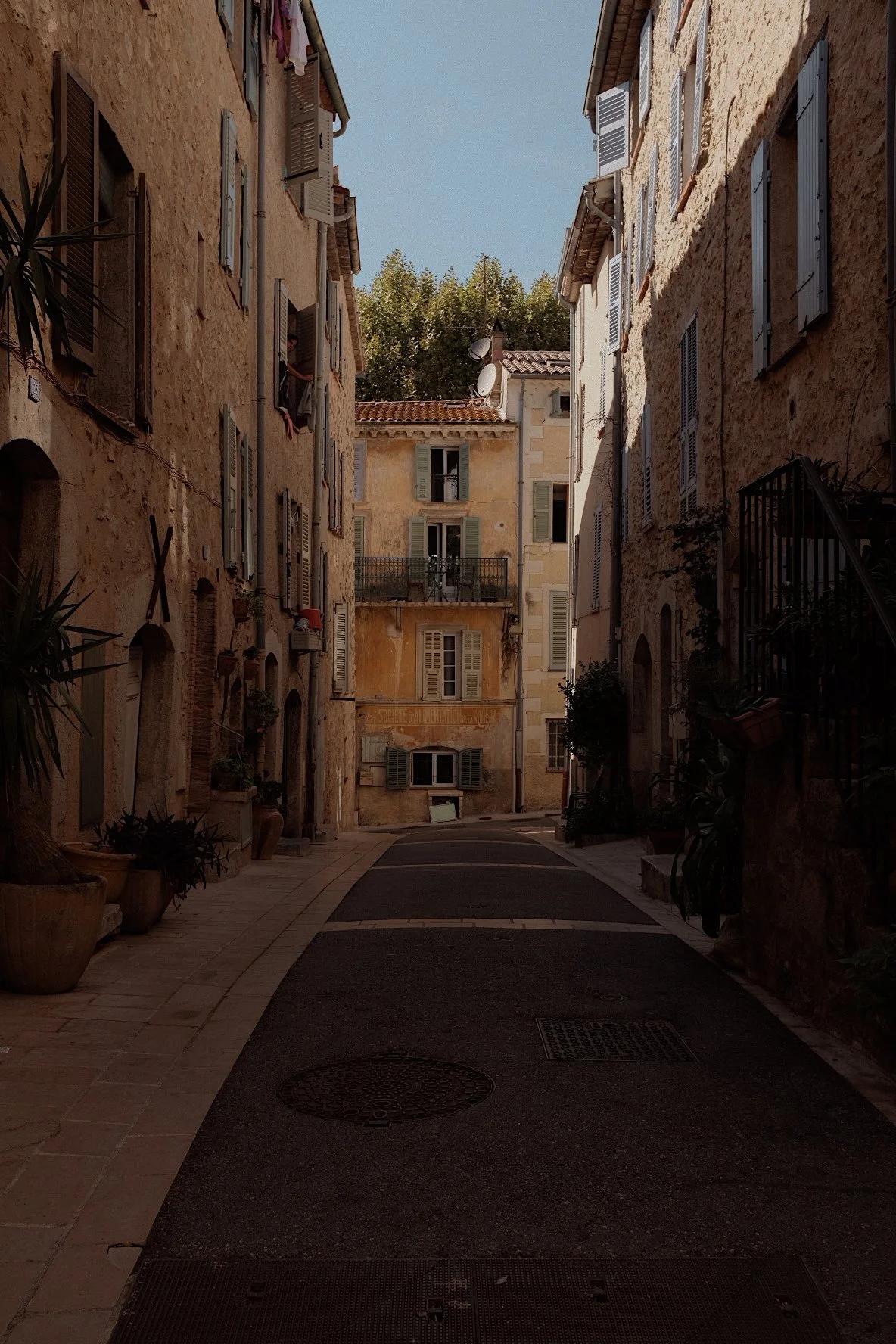 A narrow street in a Mediterranean town with old beige stone buildings on either side. The buildings have green shutters on the windows, some of which are open. Potted plants line the street, and laundry hangs from a balcony. The sky is clear, and th