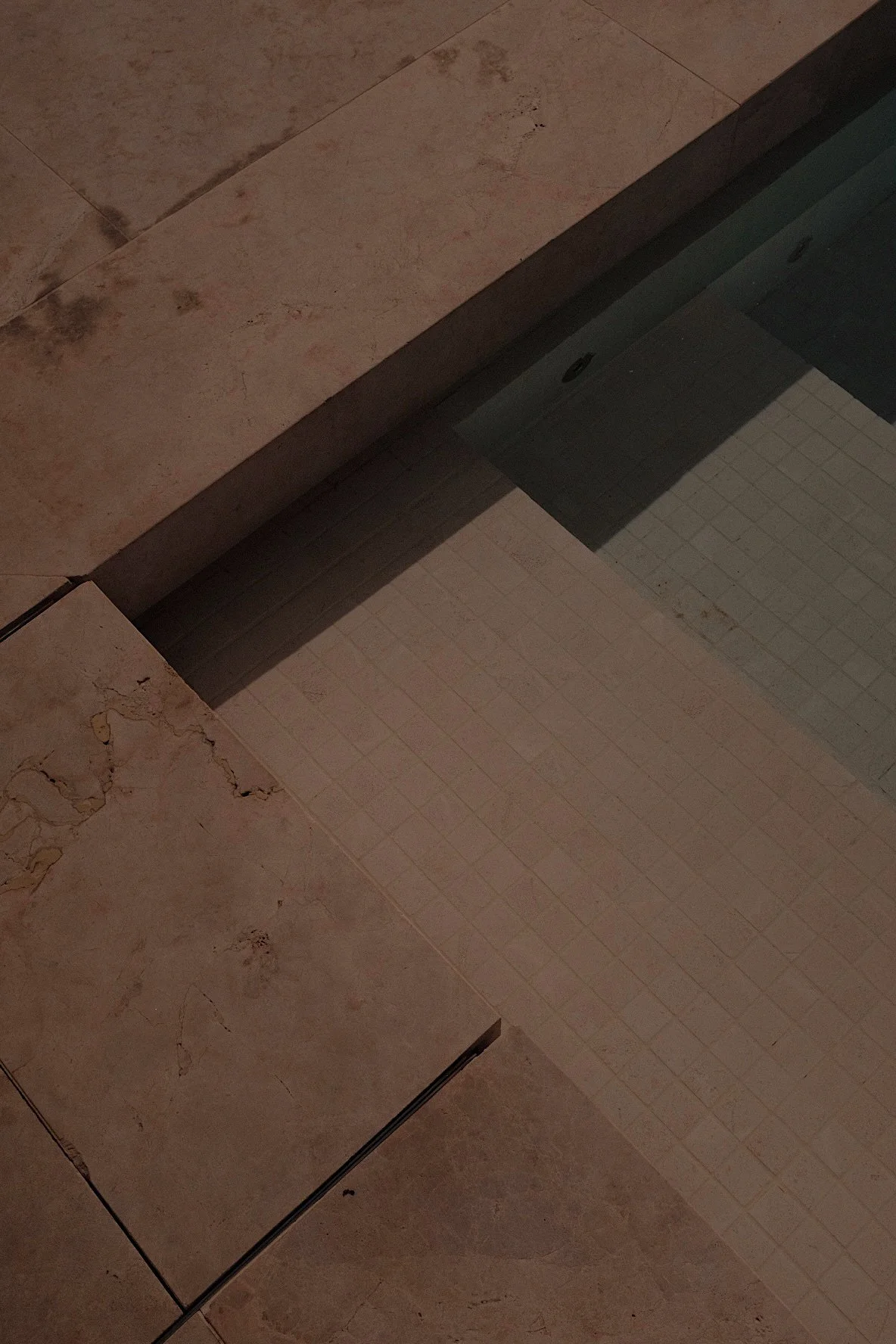 Close-up of beige tiled bathroom floor with dark stains and cracks, and part of a white tiled wall and metallic cabinet door.