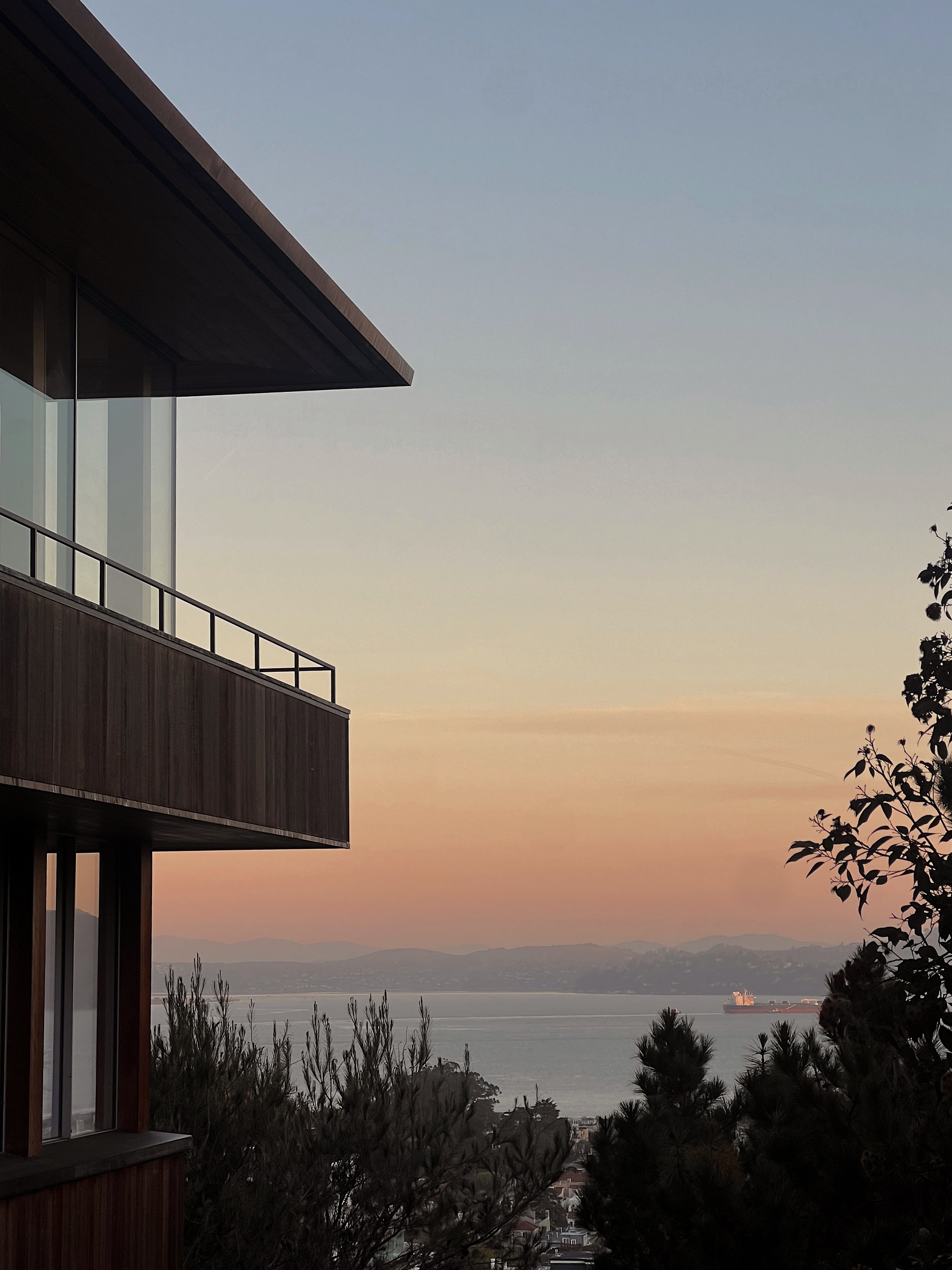 Sunset view from a modern building with glass windows, overlooking a body of water and distant hills. Trees are visible in the foreground, and a large ship is on the water. The sky is a soft gradient from blue to orange.