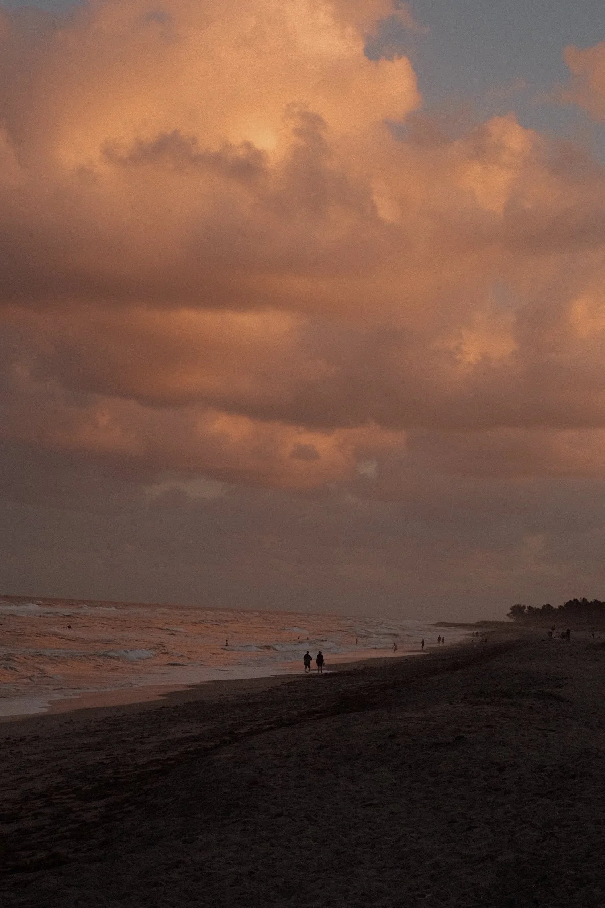 Beach scene during sunset with dark clouds overhead, waves crashing on the shore, and a few people walking along the beach.