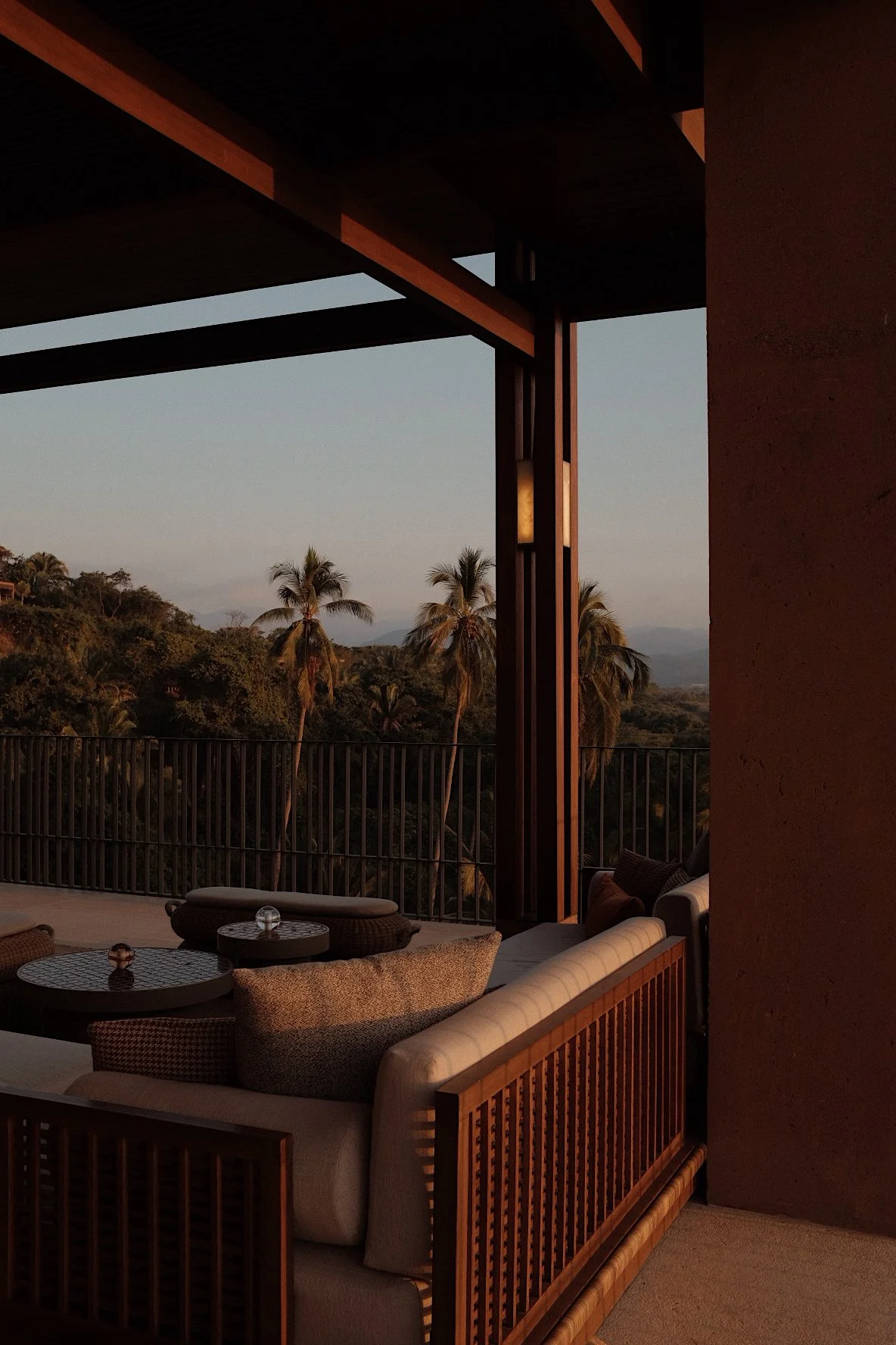 An outdoor porch or balcony with wicker furniture, including a round coffee table with glass ornaments, overlooking a landscape with palm trees and distant hills at sunset.