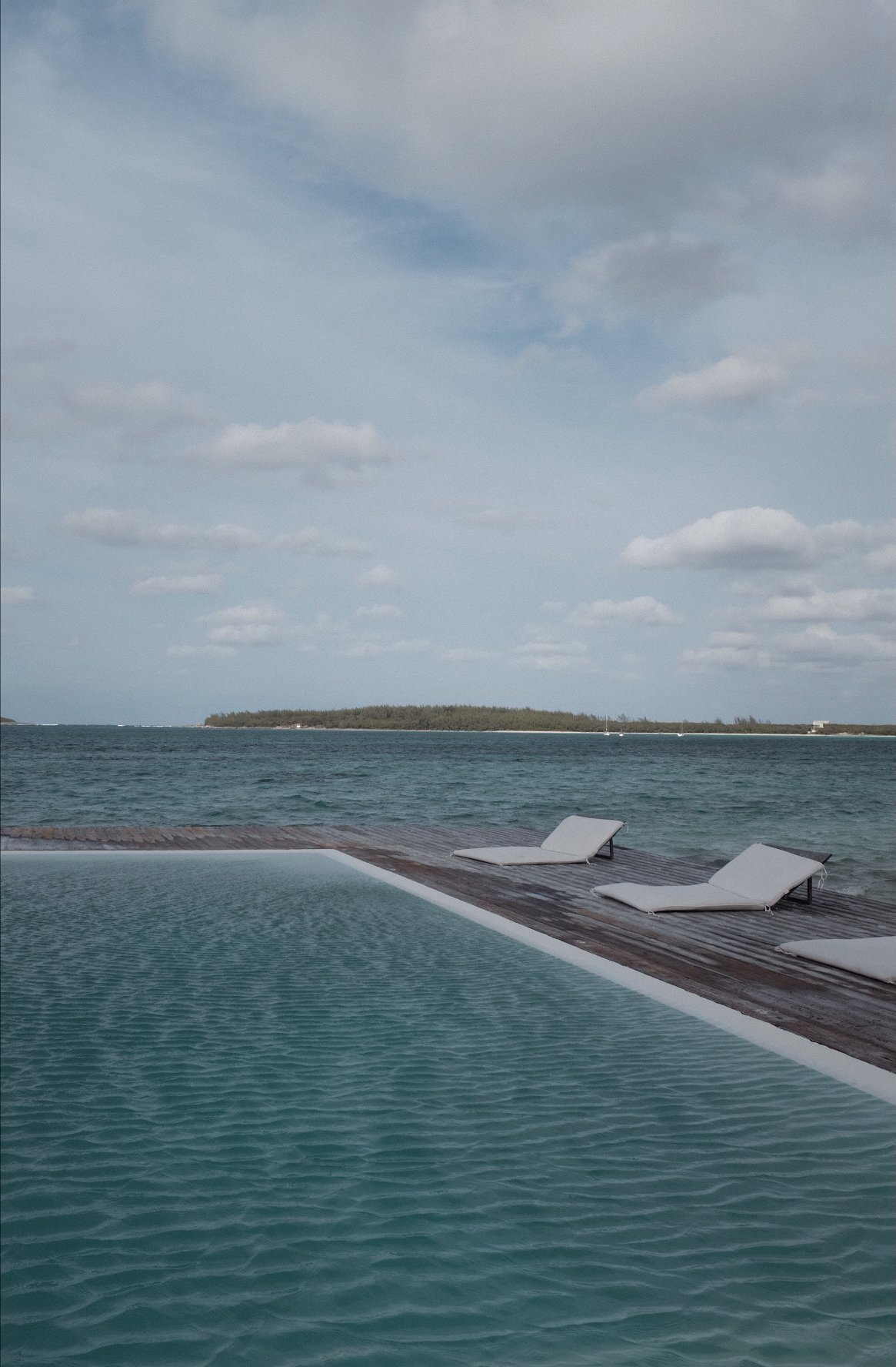 Infinity pool overlooking the ocean with two lounge chairs on a wooden deck, distant island and cloudy sky.