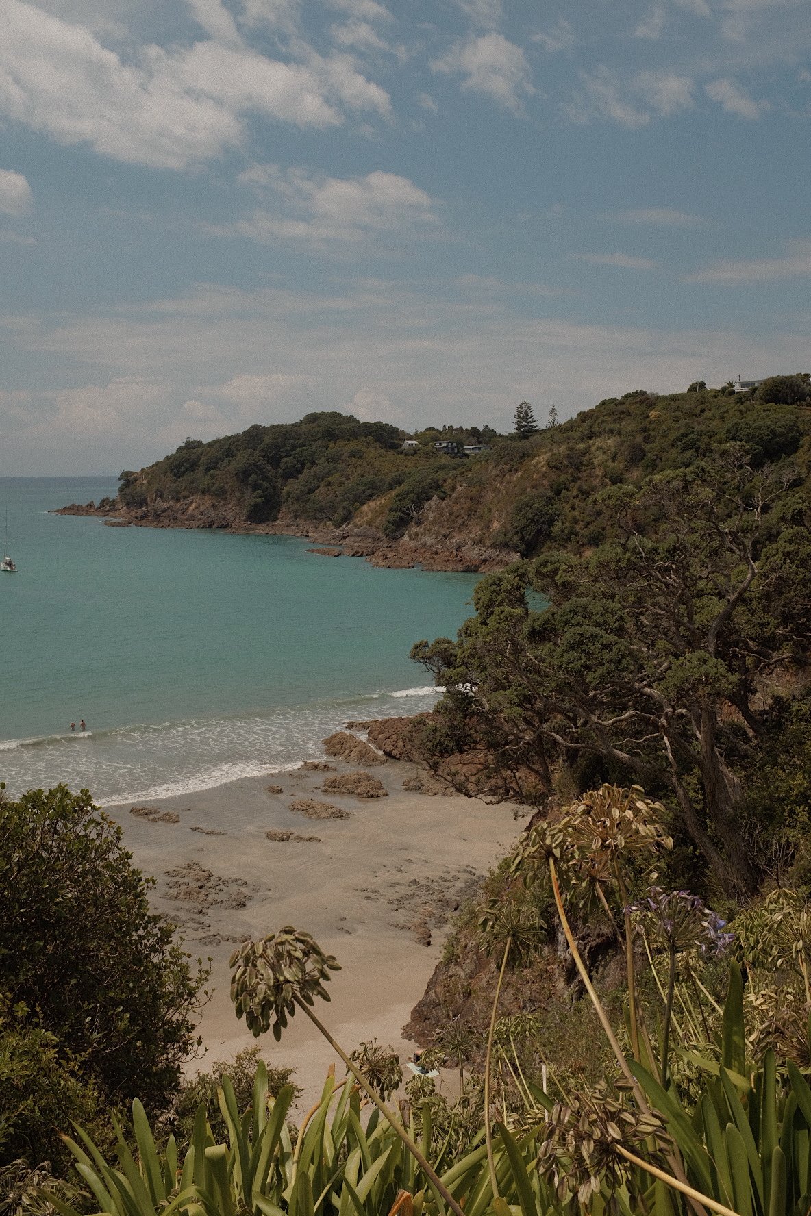 A scenic view of a coastal area with turquoise water, rocky shoreline, lush green hills, and overhanging trees. A small beach is visible with two people in the water. The sky is partly cloudy.