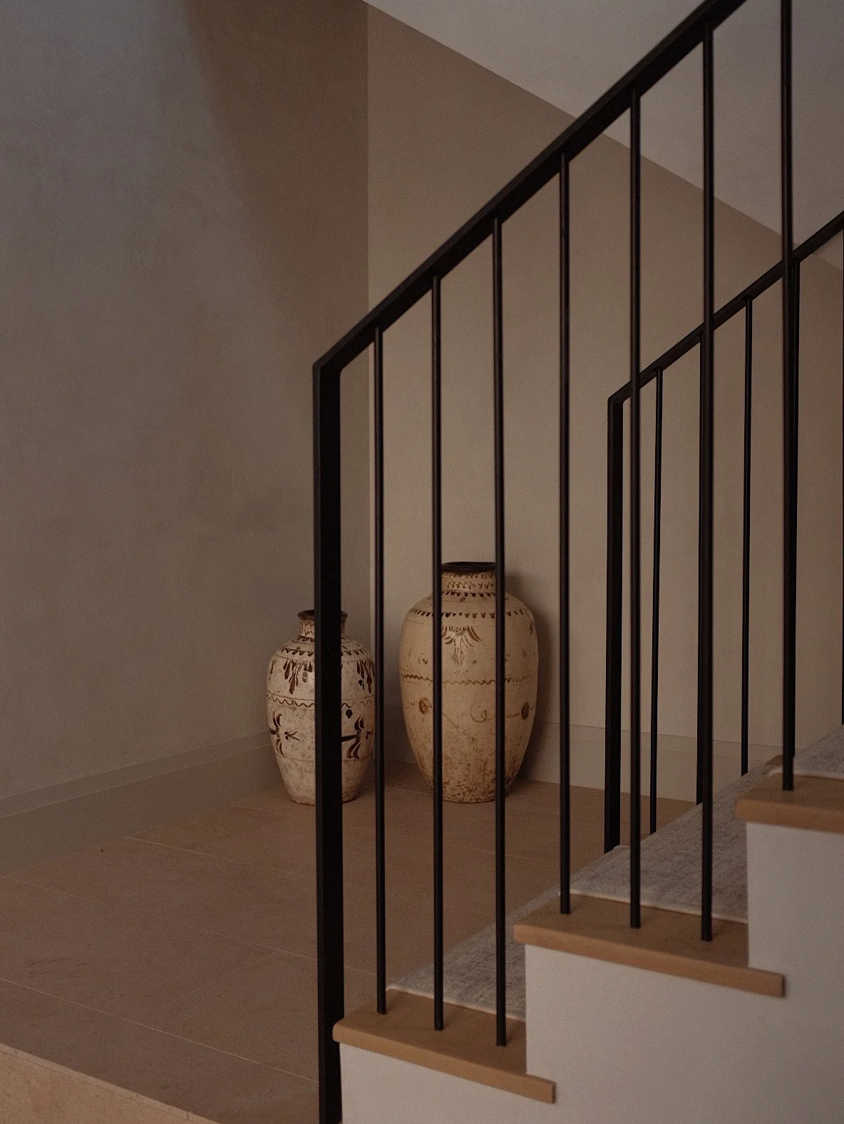 Two large decorative pottery vases with earthy patterns placed in a corner behind a black metal stair railing inside a home.