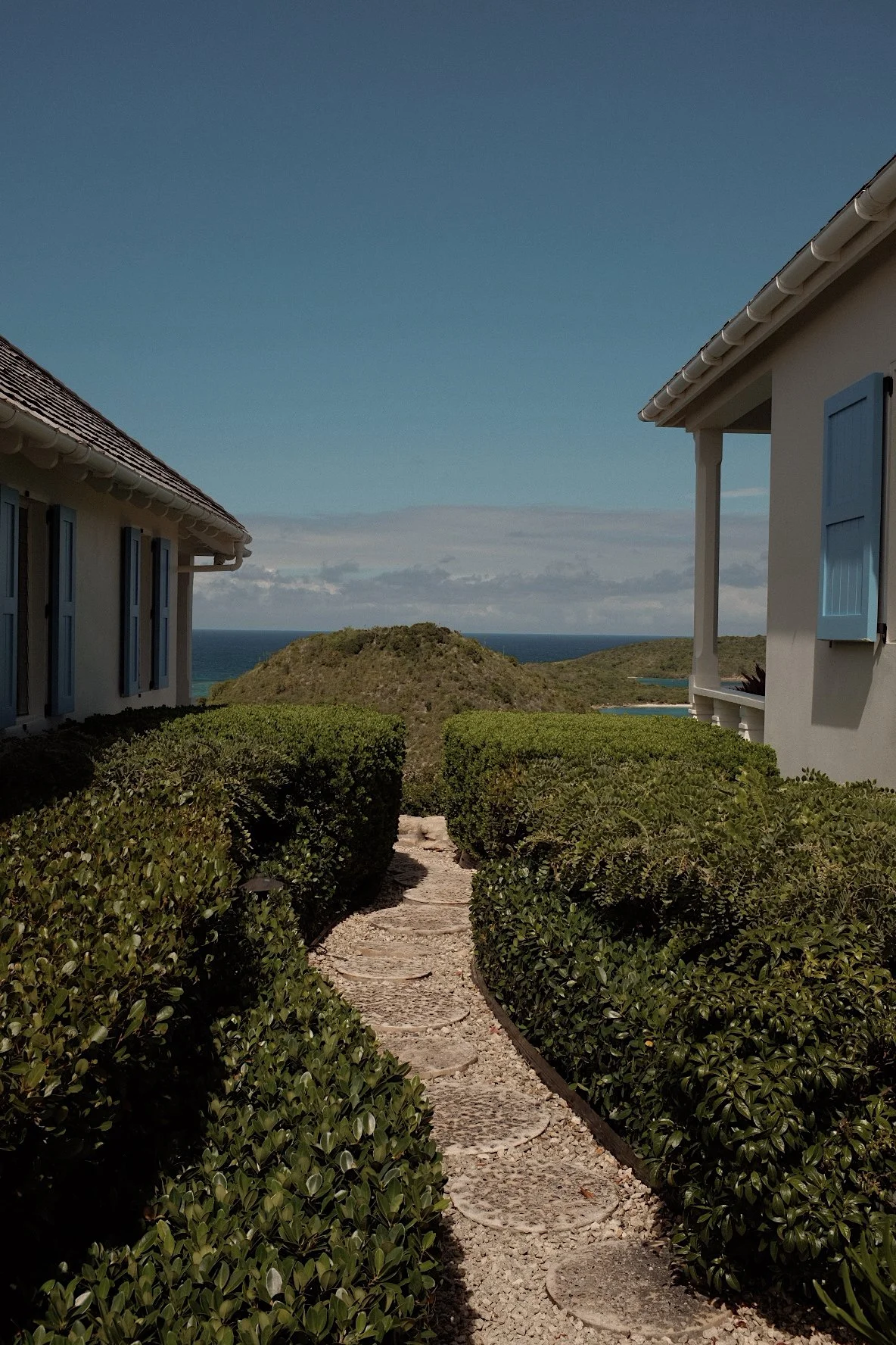Stone pathway lined with trimmed greenery leading to distant ocean view, flanked by two houses with blue shutters.