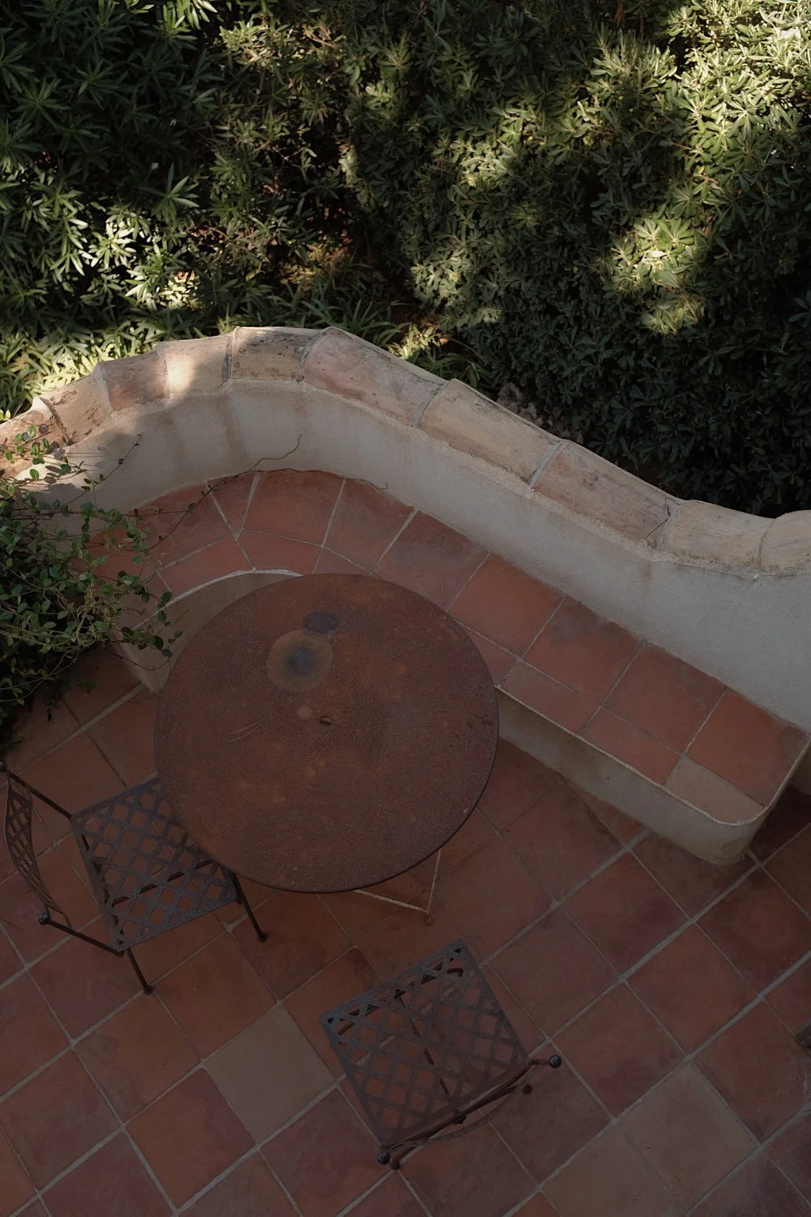 A small round rusted metal table with two matching metal chairs on a tiled patio. A curved brick and stucco wall borders a garden with green foliage and shadows.