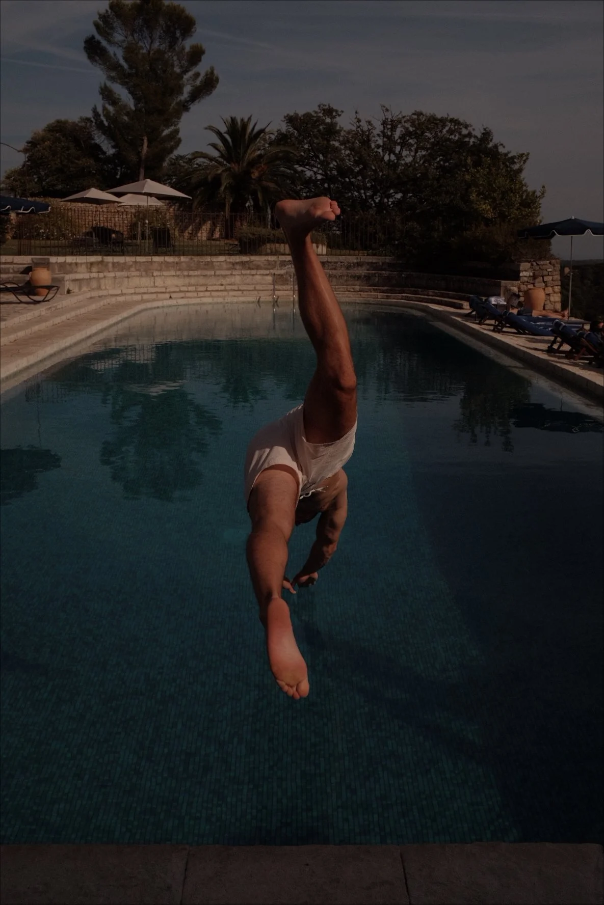 A person performing a handstand on the edge of a swimming pool during sunset, with trees, umbrellas, and lounge chairs in the background.