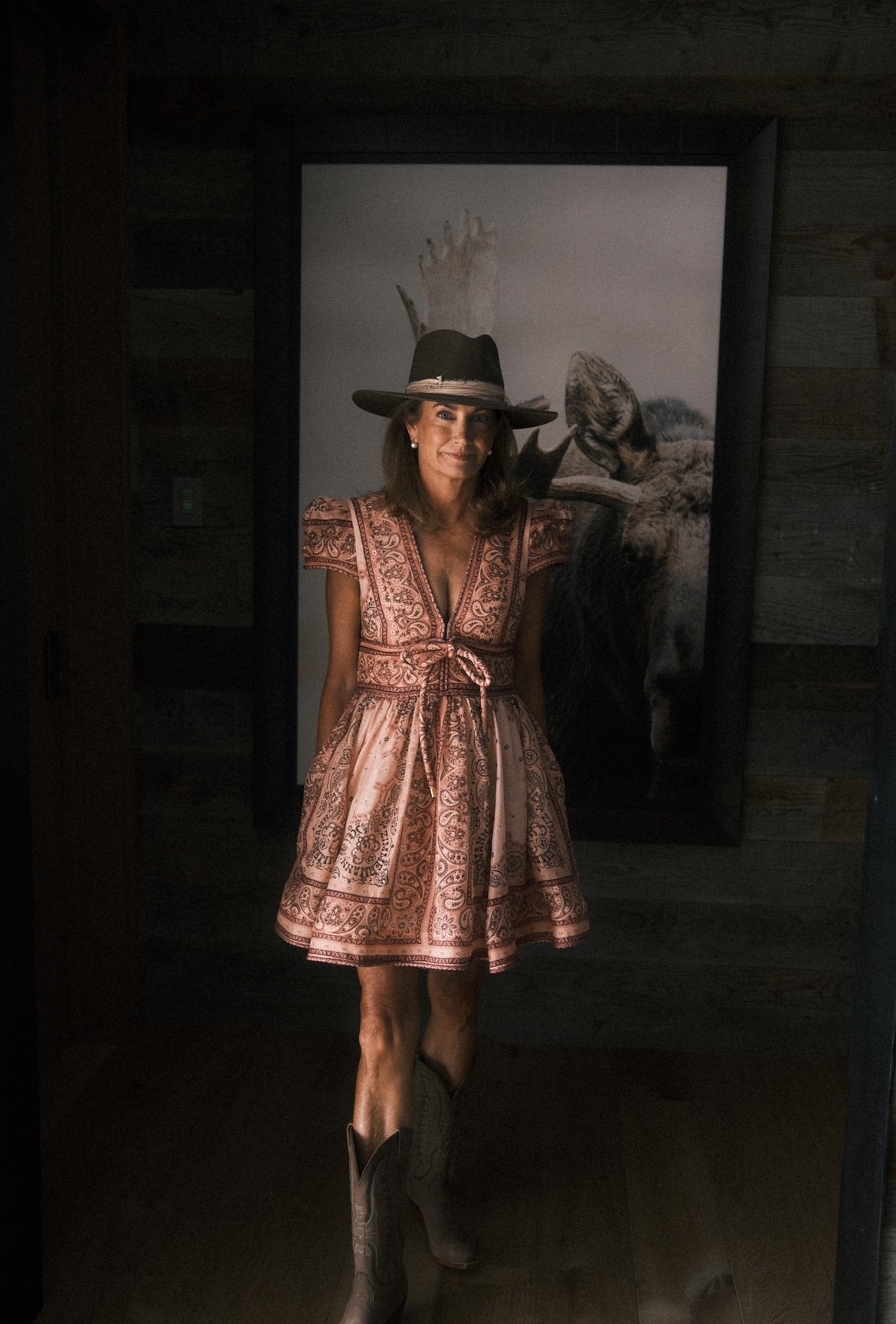 Woman wearing a hat, patterned dress, and cowboy boots standing indoors with an animal artwork in the background.