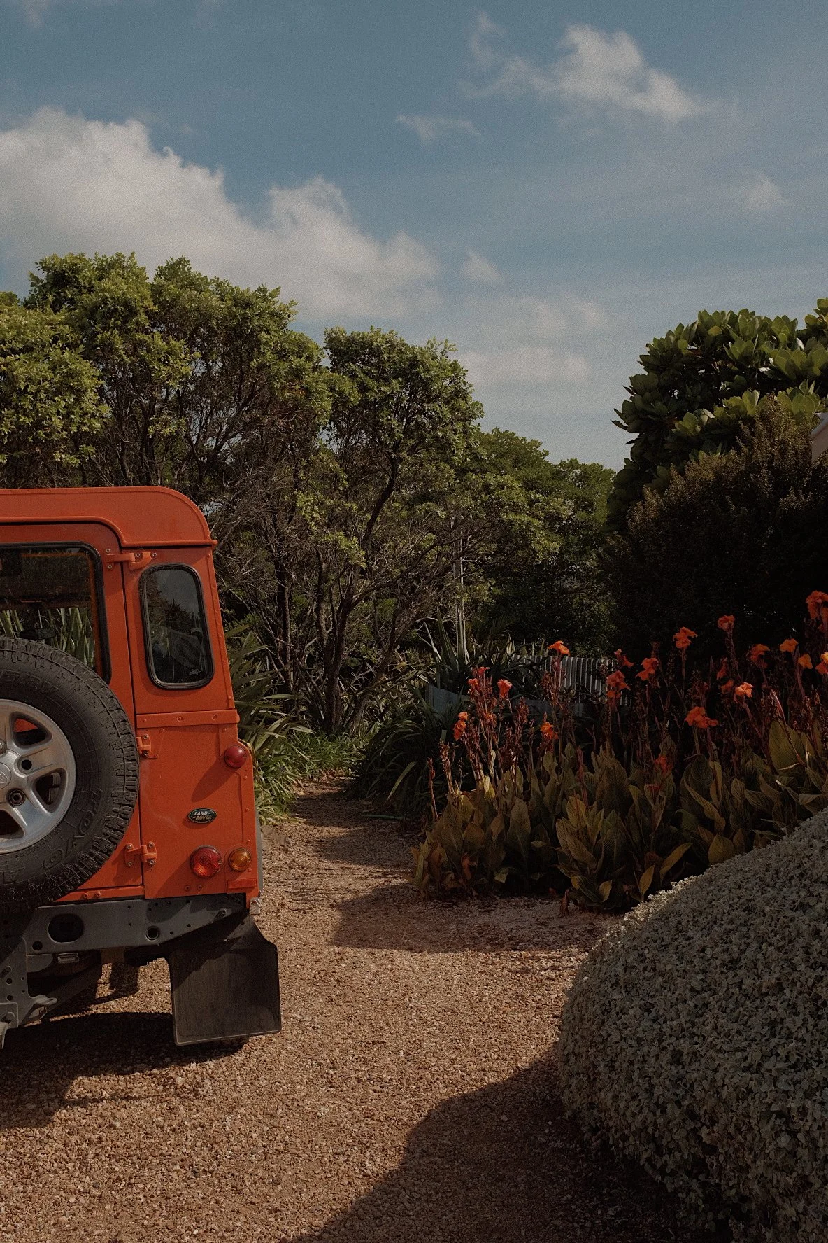 Orange vehicle parked on a gravel path surrounded by lush green trees and vibrant red flowers under a blue sky.