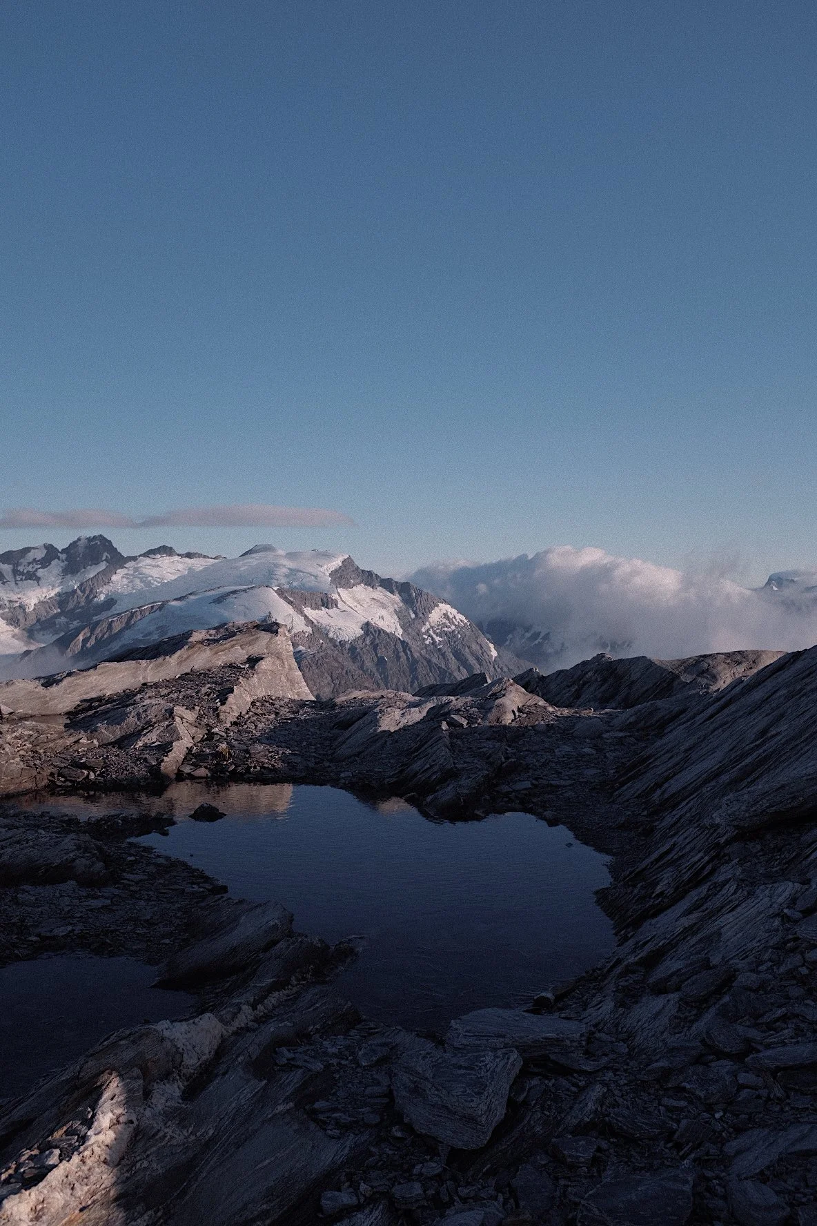 Snowy mountain landscape with rocky terrain and a small reflecting pond under a clear blue sky.