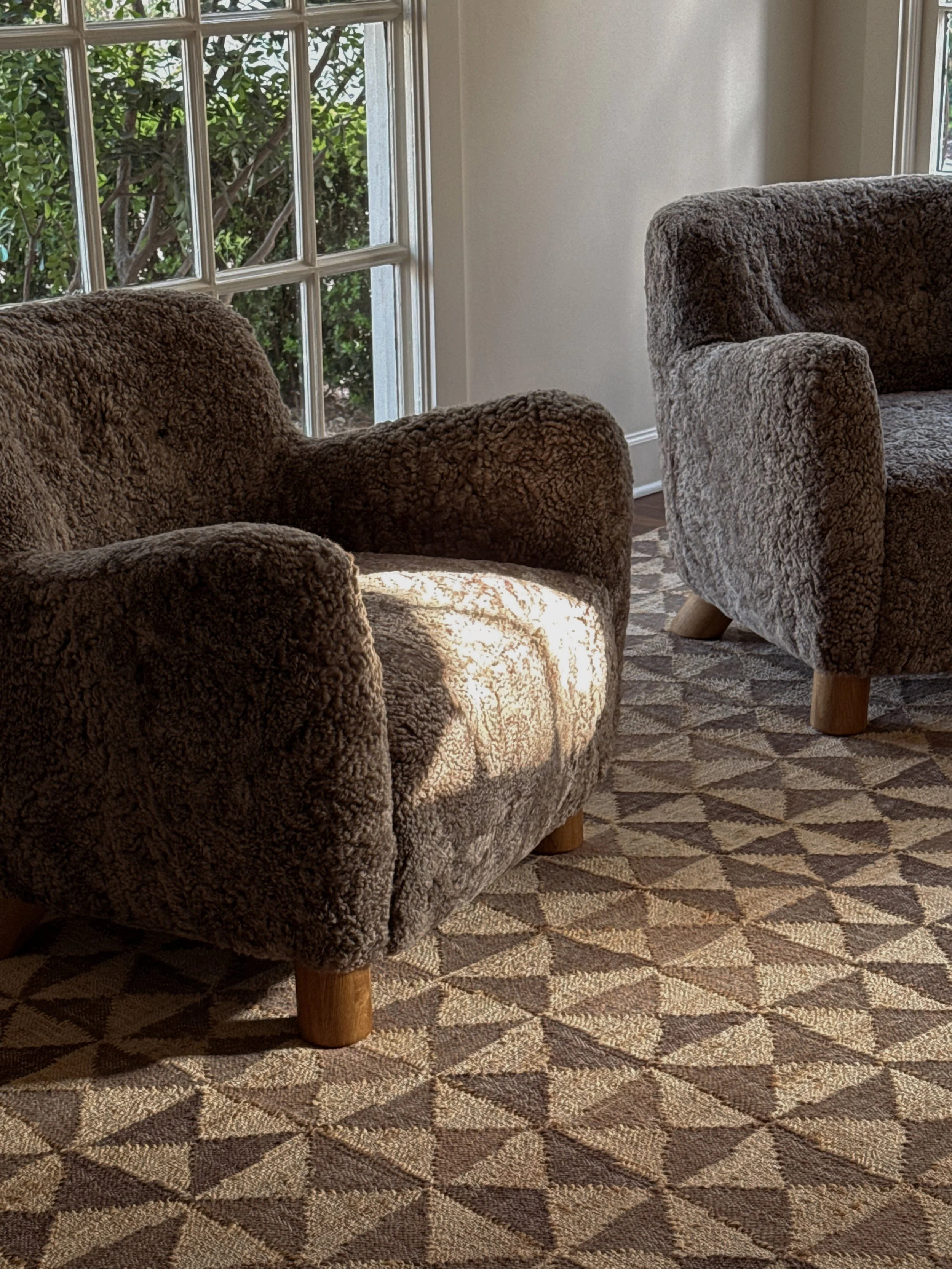 Two plush armchairs with brown and grey fuzzy fabric, positioned in a sunlit living room next to a patterned rug, with two large windows showing green foliage outside.