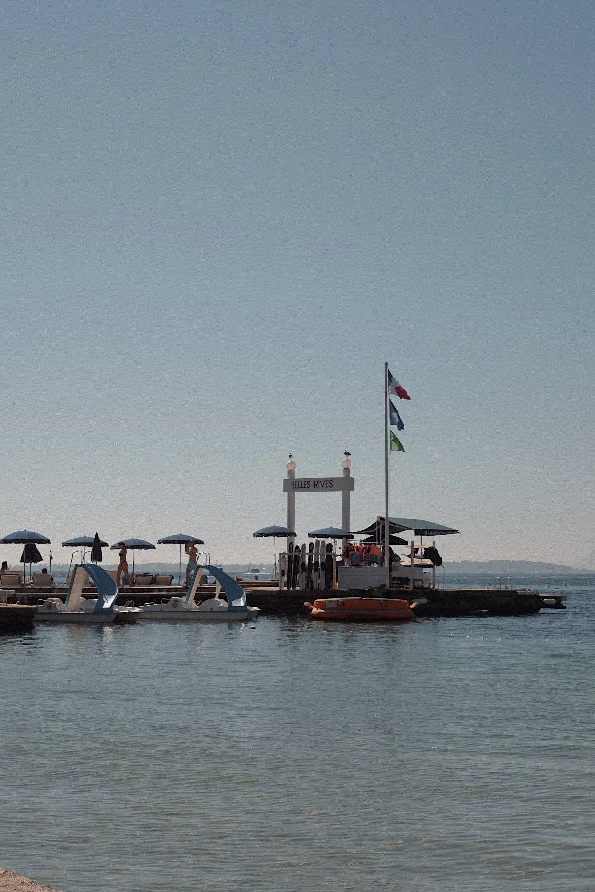 A beach scene at Belles Rives with a pier, umbrellas, and boats on the water under a clear sky.