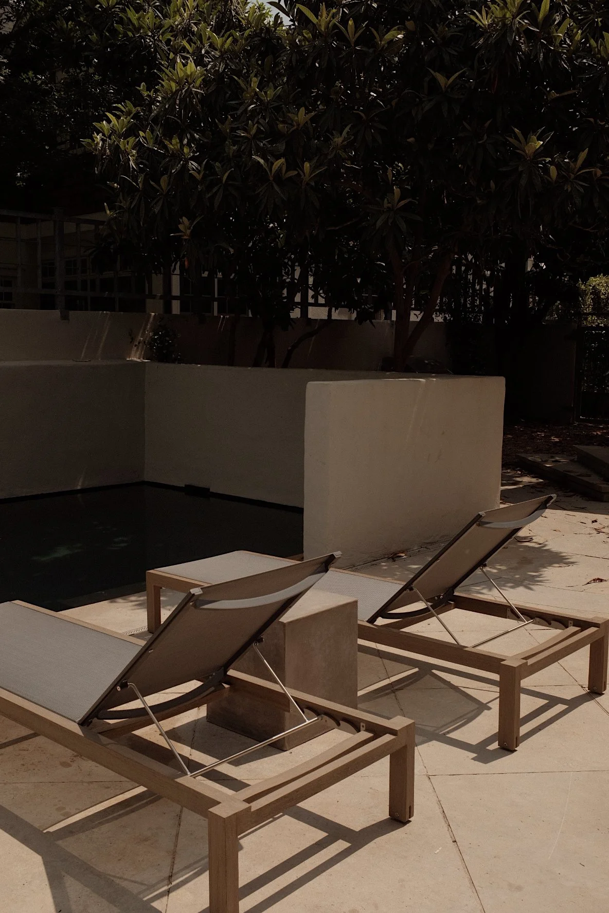 Two wooden lounge chairs with beige fabric are on the patio next to a small pool, with a low wall, trees, and bushes in the background during daytime.