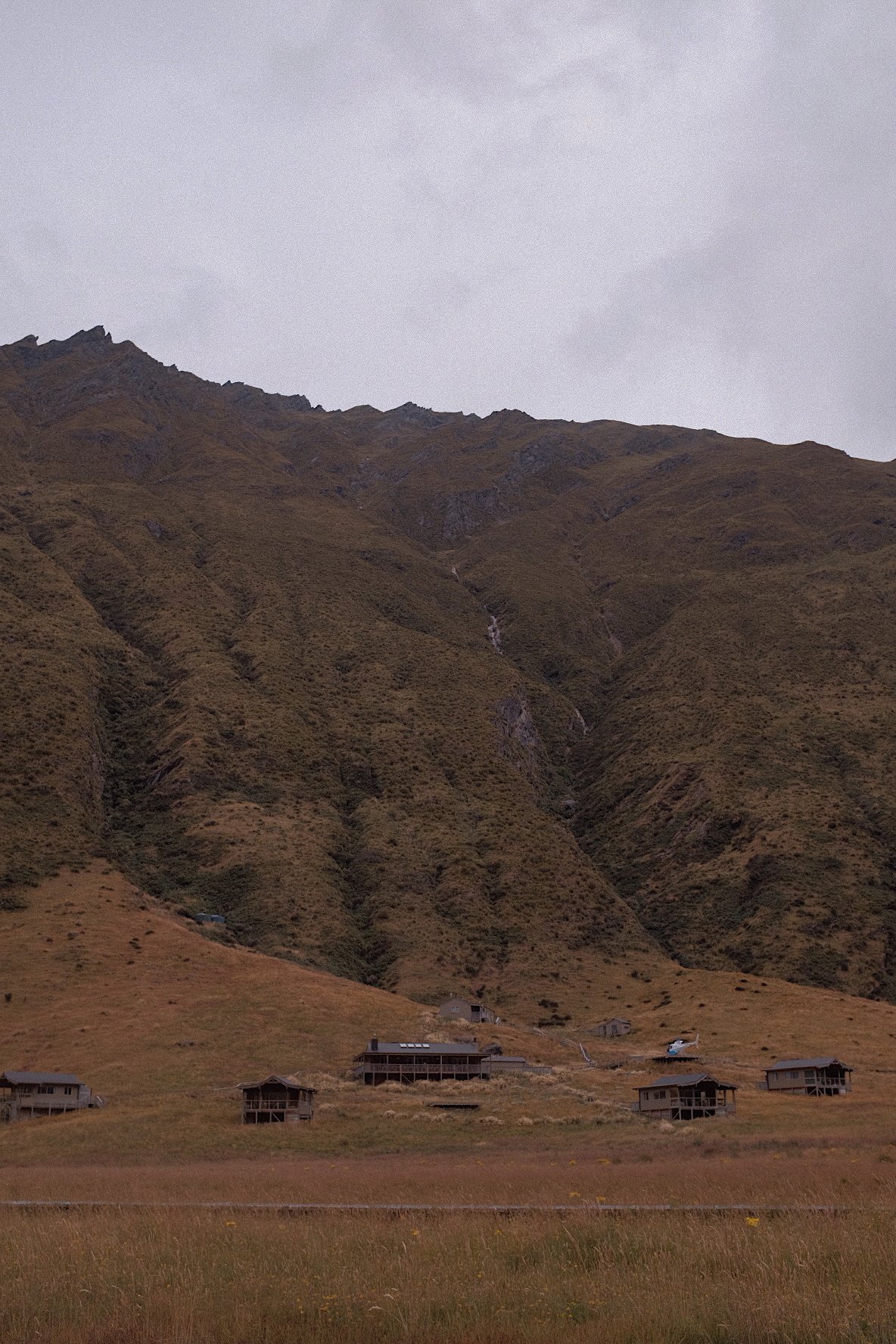 Houses on grassy hills with mountain background under cloudy sky.
