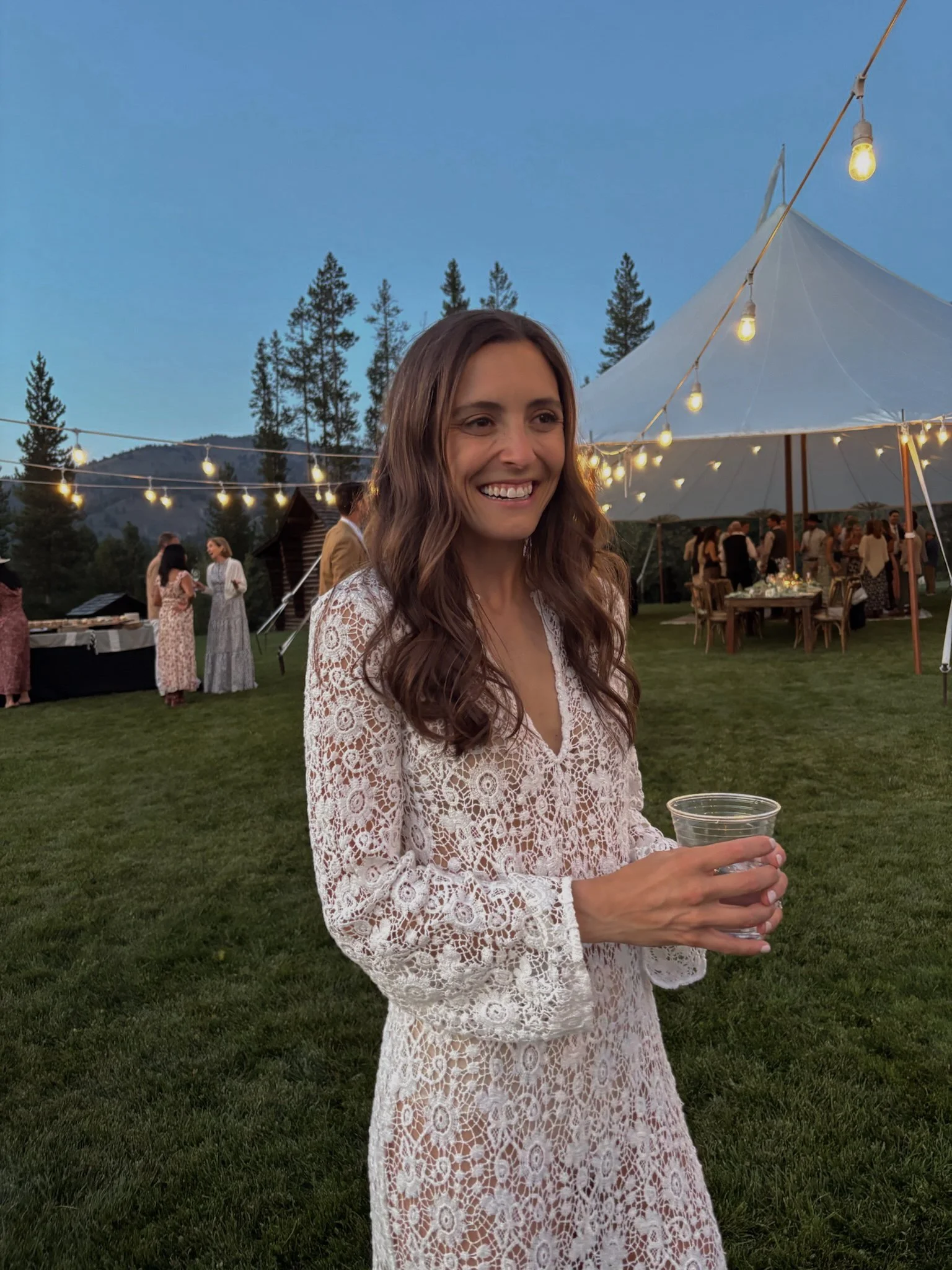 A woman with long wavy brown hair wearing a white crocheted lace dress, smiling and holding a clear plastic cup, at an outdoor evening event with string lights, a white tent, and other people in the background, surrounded by trees and mountains.