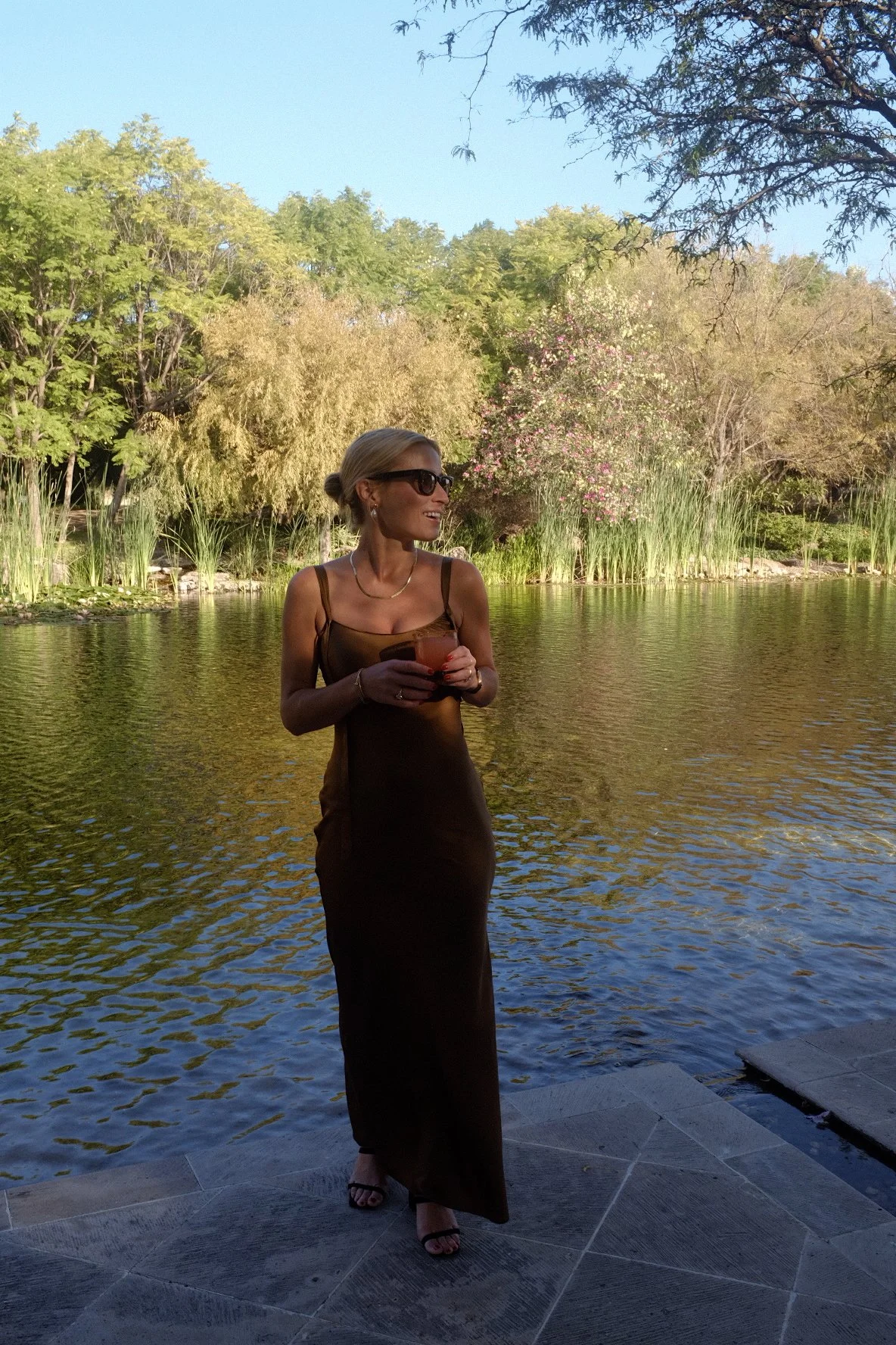 Woman in a long dress standing near a pond, holding a drink, with trees and plants in the background.