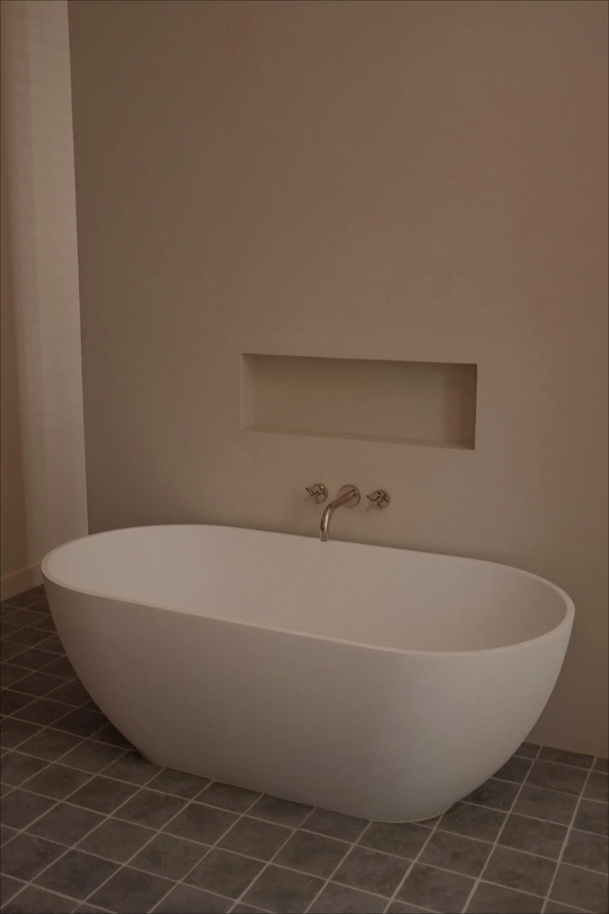 Modern freestanding white bathtub with a metal faucet on a tiled bathroom floor, with a recessed wall niche behind it.