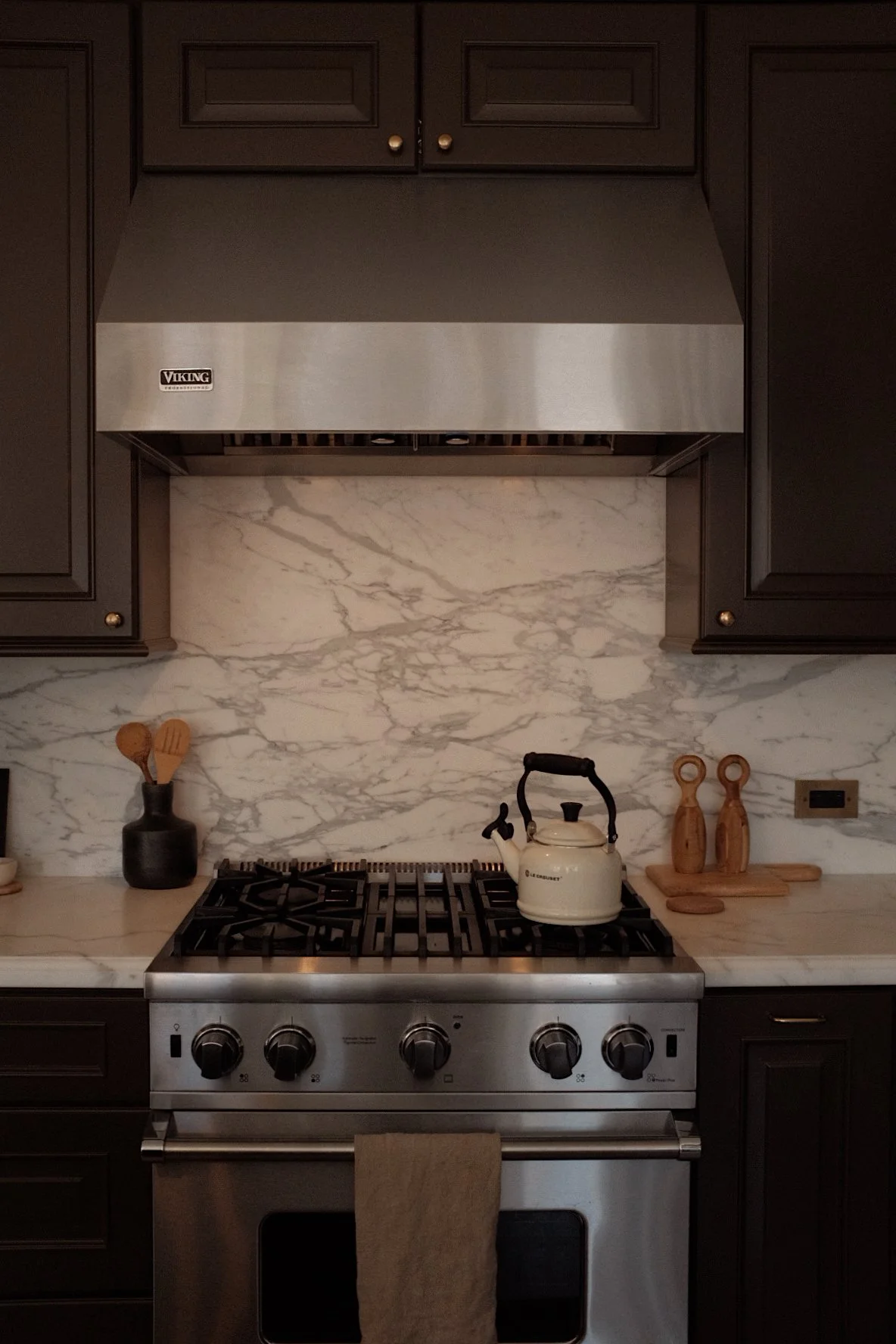 Kitchen stove with a white kettle on top, black cabinetry, a marble backsplash, and wooden utensils on the counter.