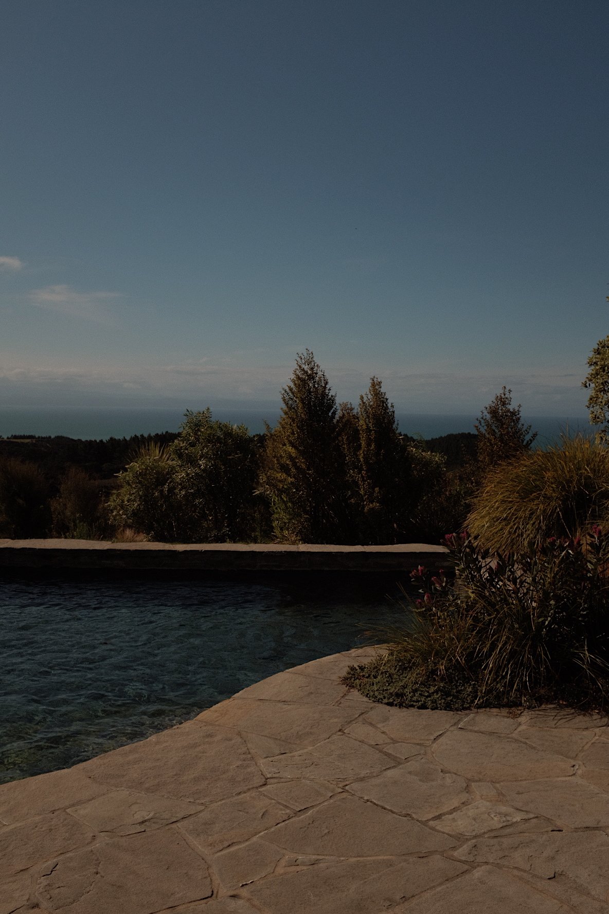 Outdoor stone patio with swimming pool and scenic view of trees and ocean under clear blue sky.