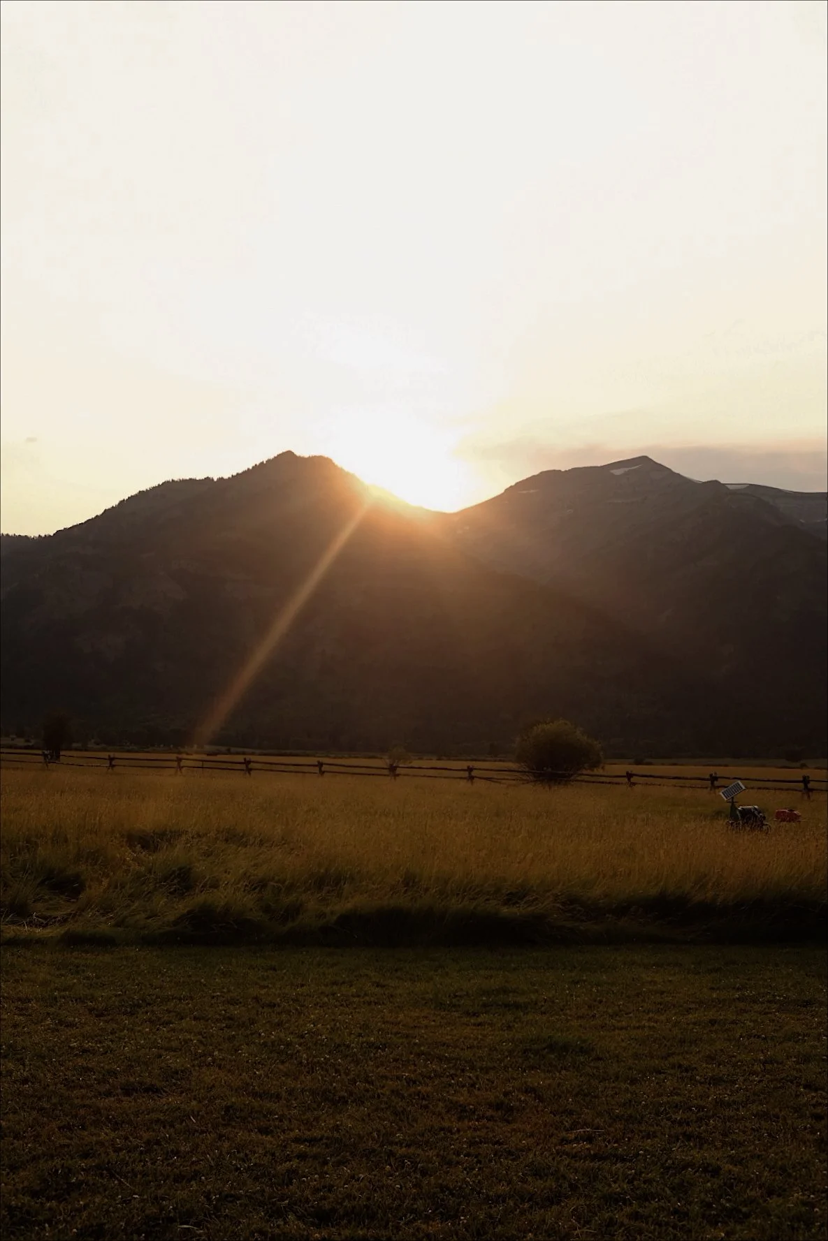 Sunset over mountains with a grassy field in the foreground and a wooden fence. The sun is partially behind the mountain, creating a bright glare and lens flare.