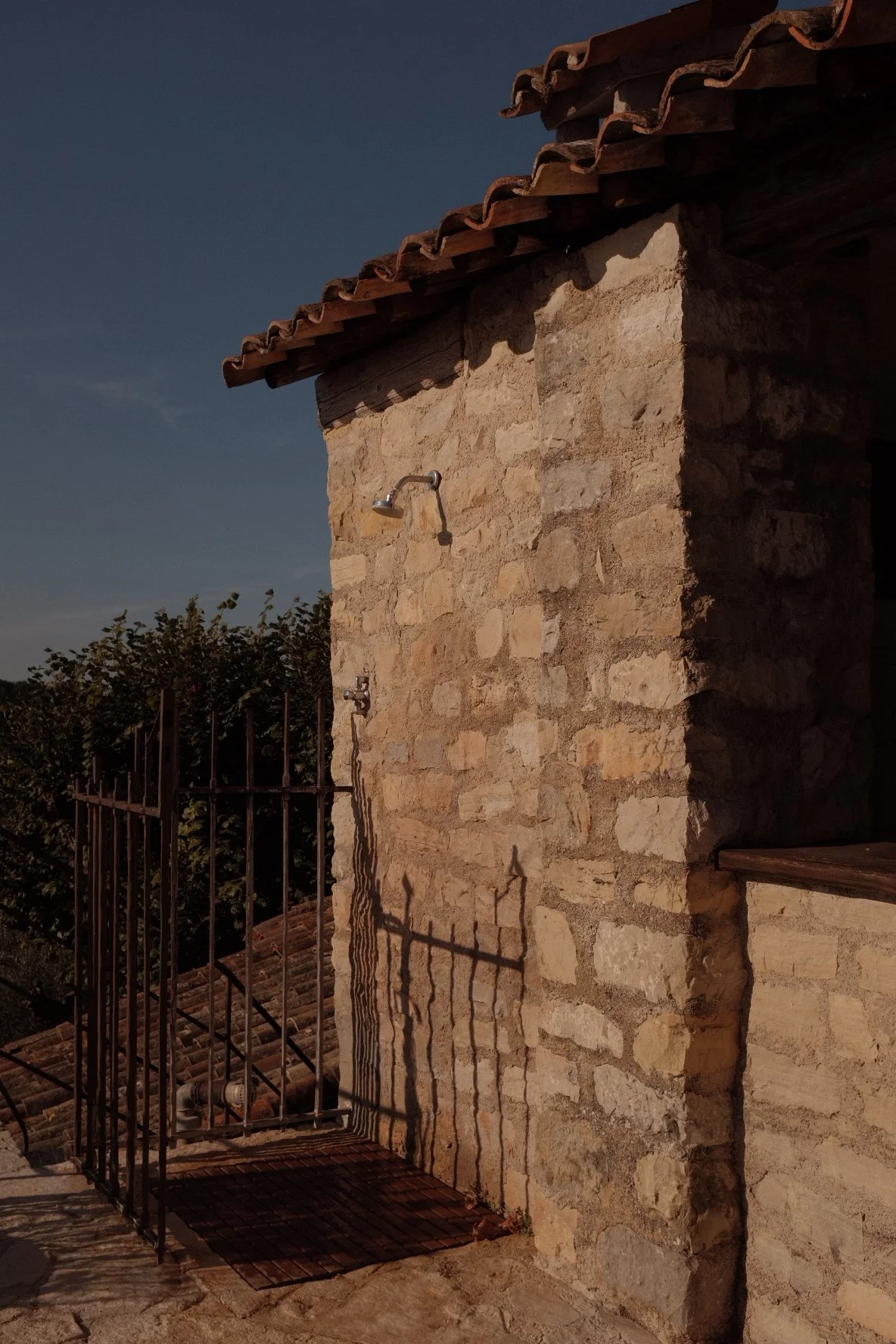 Stone building with a tiled roof, outdoor shower, and metal gate cast shadows in the sunlight.