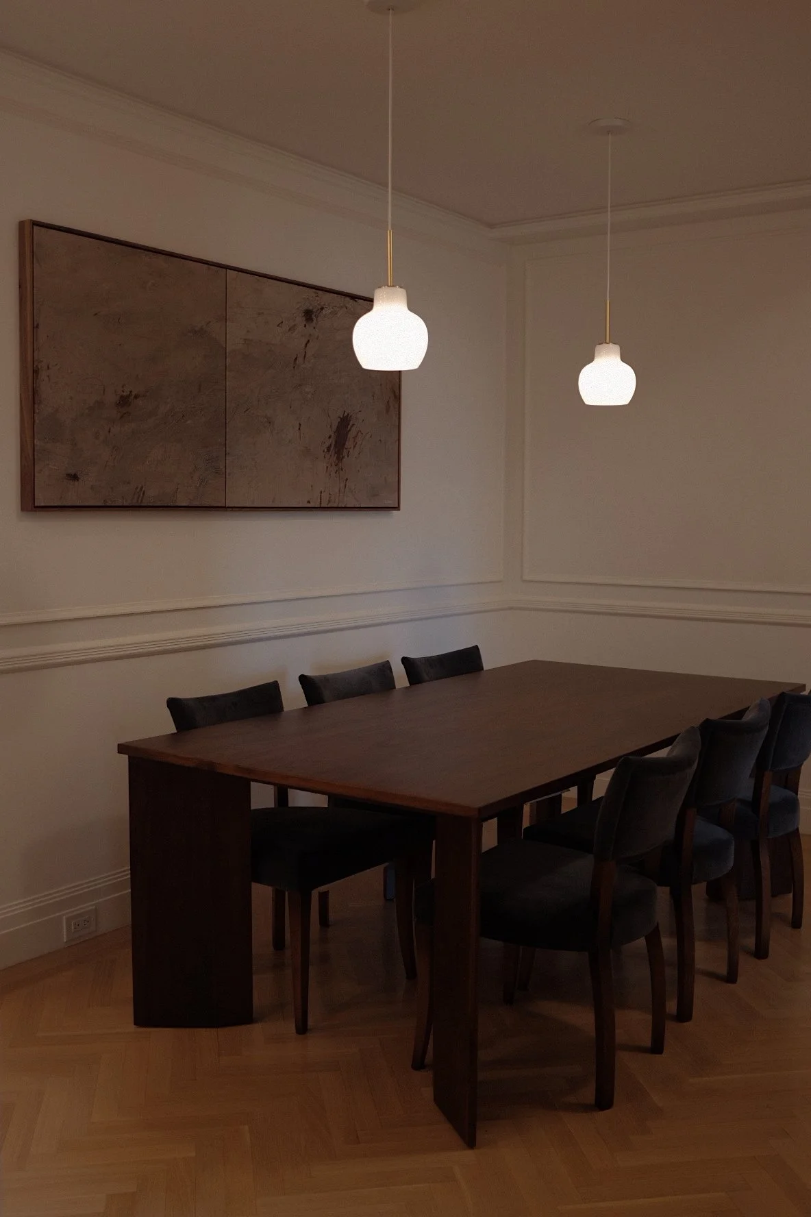 Empty dining room with a large wooden table surrounded by black chairs, two pendant lights hanging from the ceiling, a framed abstract painting on the wall, and hardwood floors.