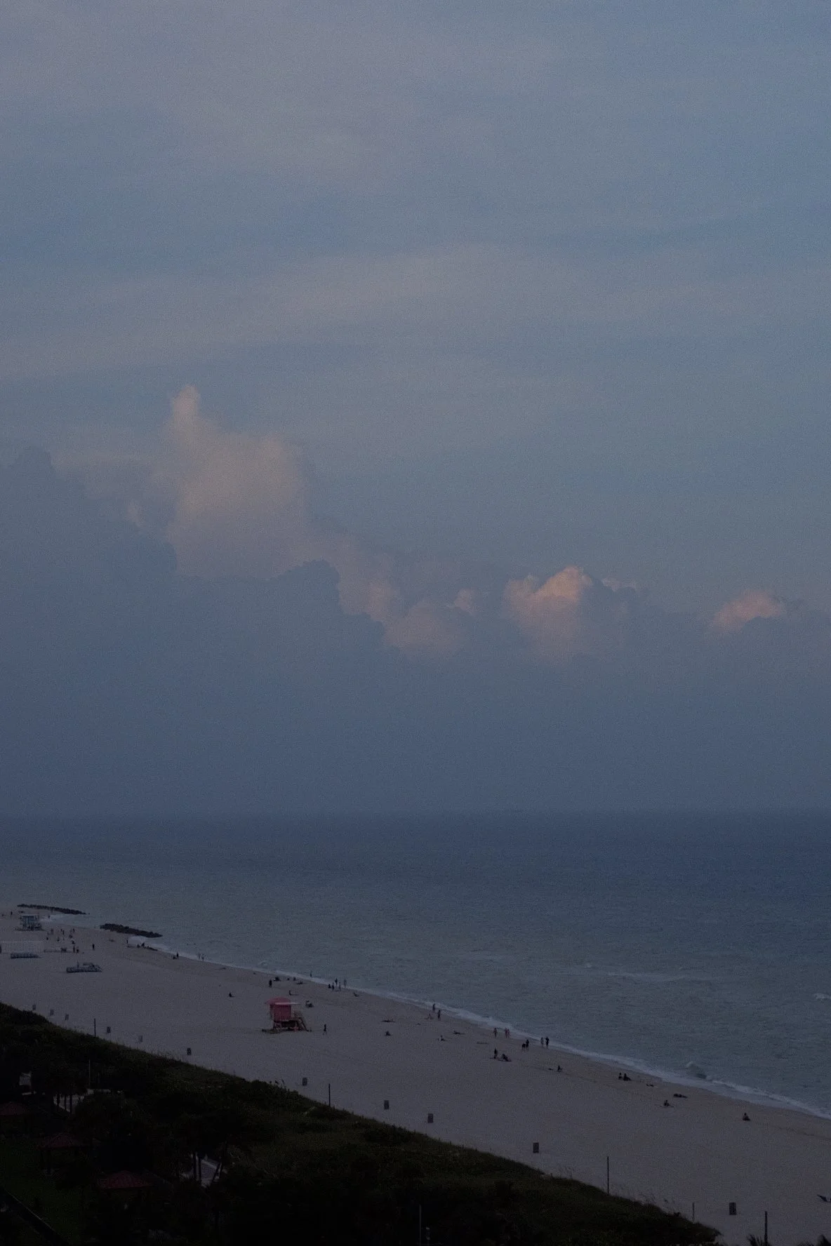 Sandy beach with a lifeguard tower, ocean waves, and cloudy sky at dusk, people walking and relaxing on the shore.