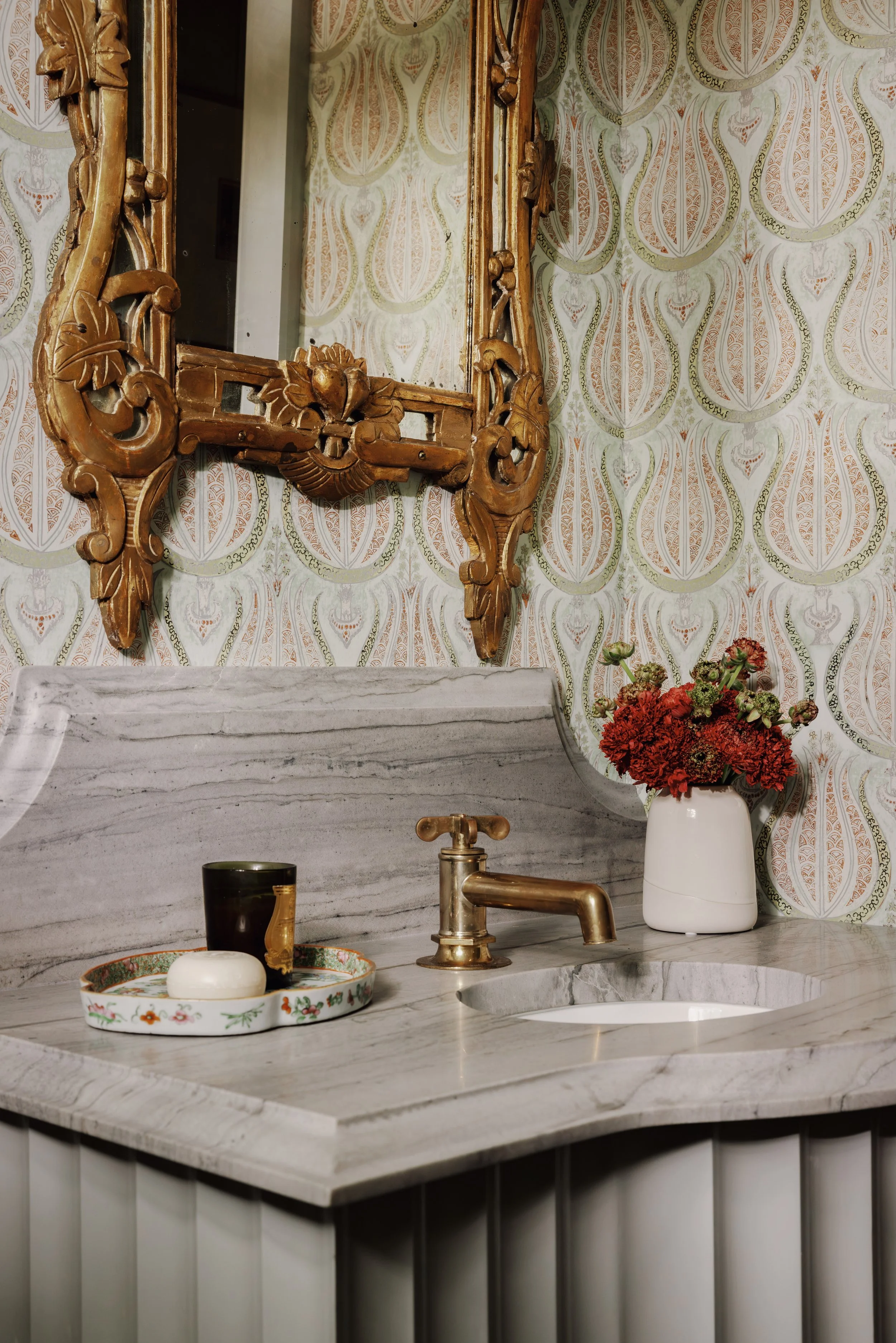 A decorated marble kitchen countertop with a floral patterned plate holding a white soap or paper towel dispenser. A dark cup, a gold faucet, and a white vase with red flowers are also on the counter. Behind the counter, there is a patterned wallpape