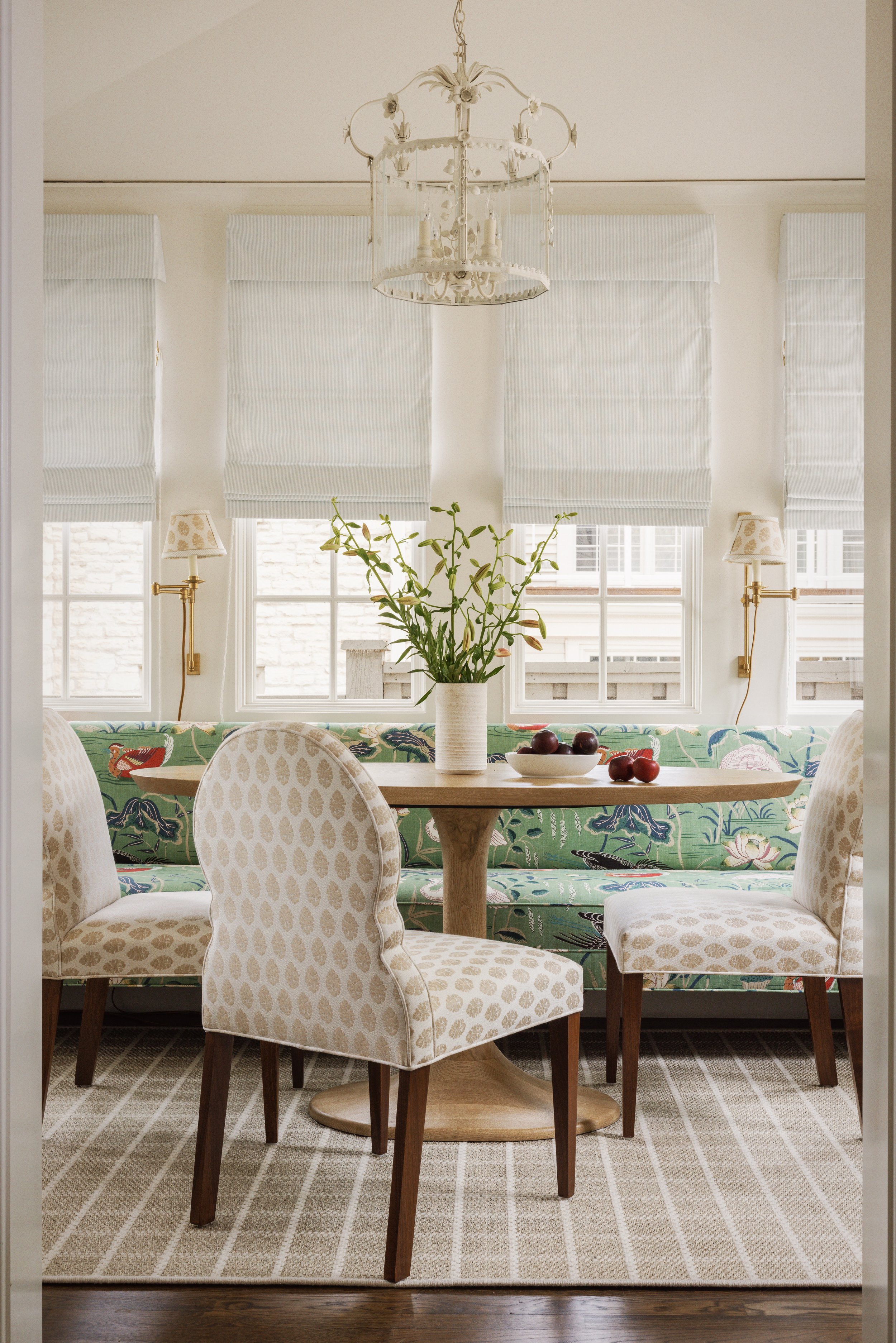 A dining area with a round wooden table, surrounded by chairs with beige upholstered fabric and dark wood legs. On the table is a white vase with green leafy plants and a bowl of red apples. Behind the table, there is a floral patterned upholstered b