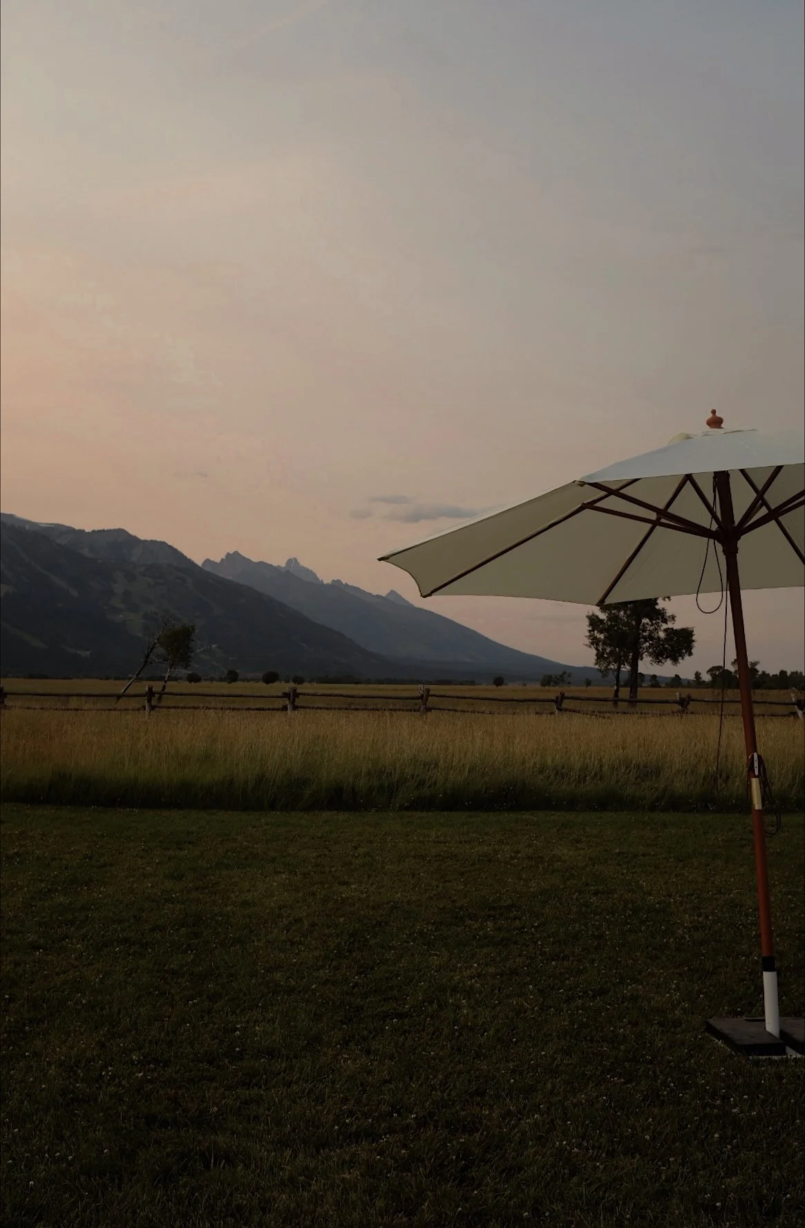 A landscape scene at sunset with mountains in the background, a grassy field, trees, a wooden fence, and a white patio umbrella in the foreground.