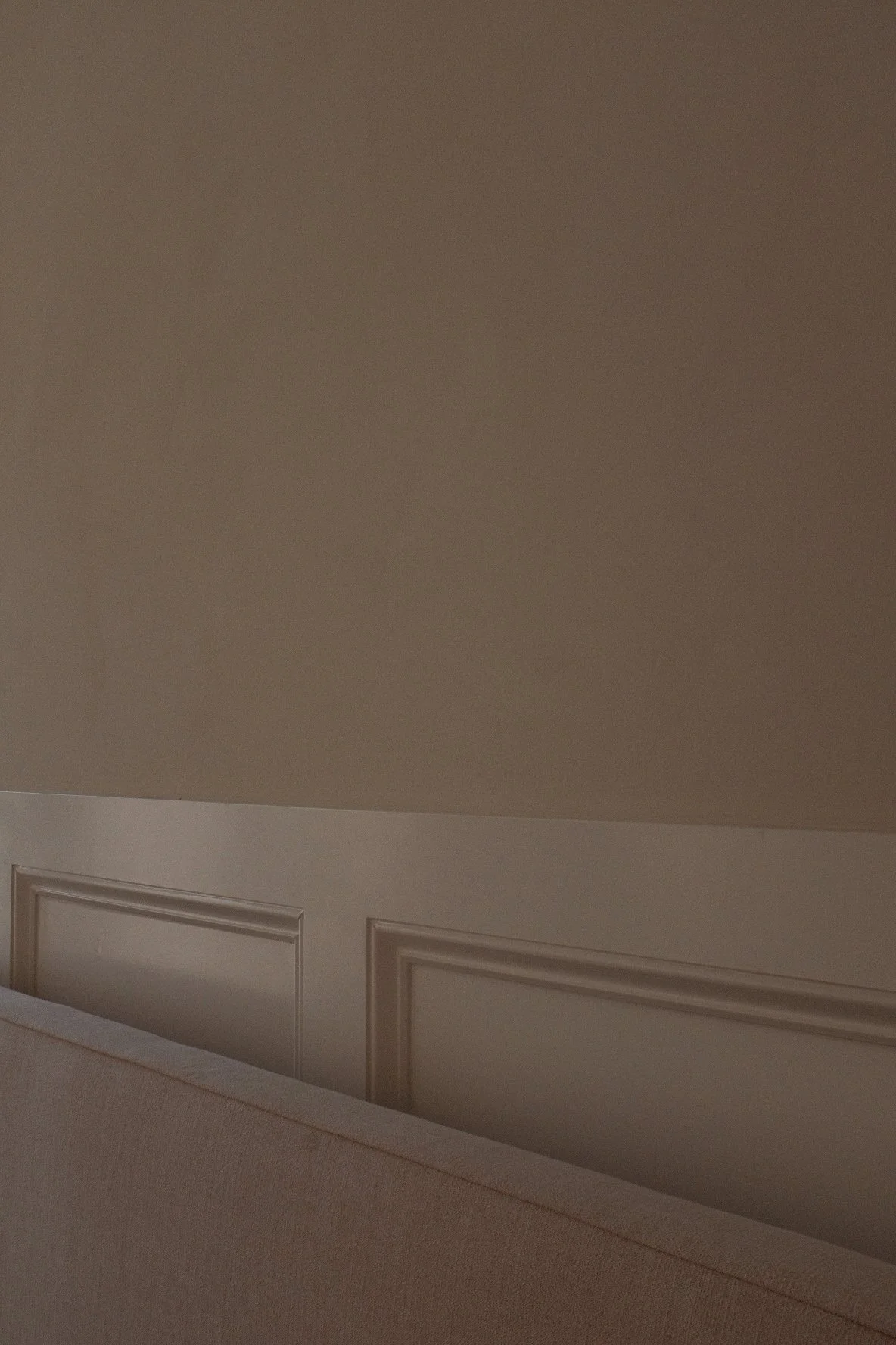 Part of a bedroom with a beige wall and white wainscoting. A beige bed with a slightly textured fabric is visible at the bottom of the image.