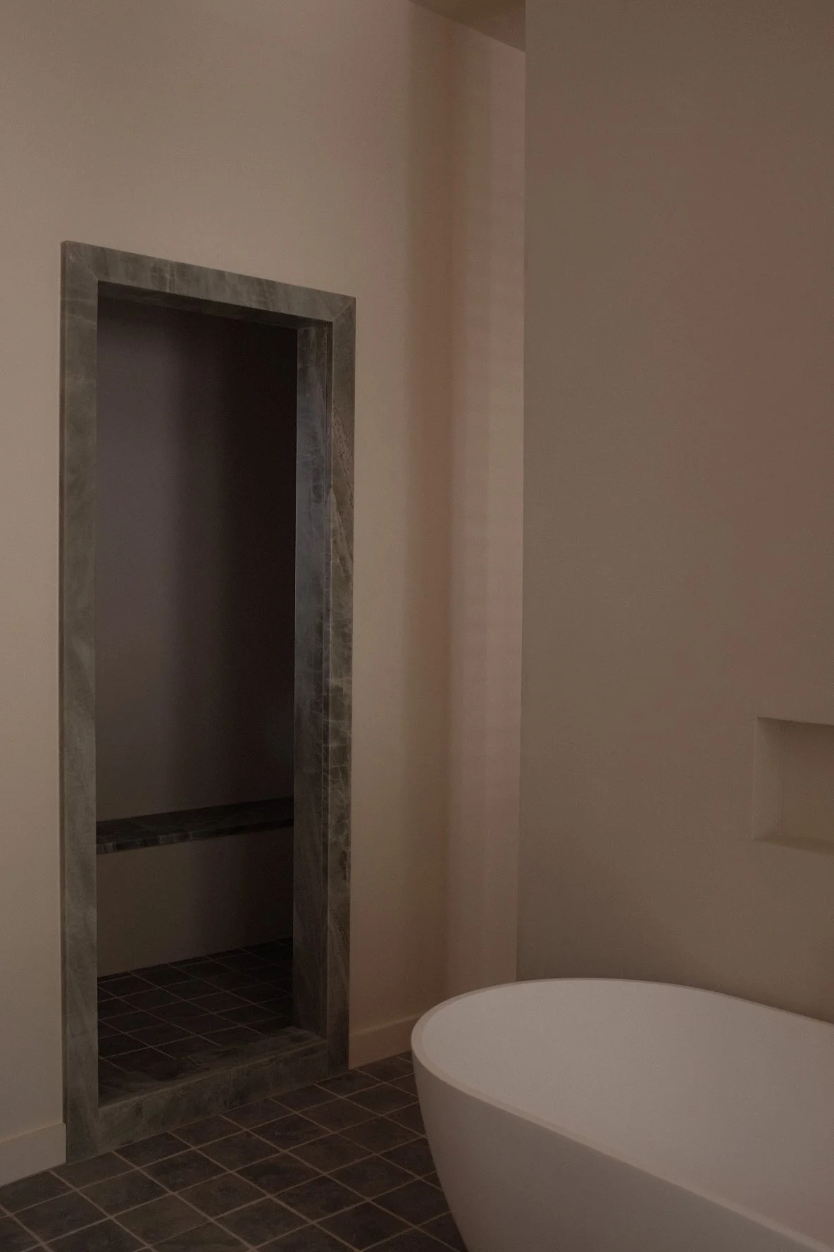 Part of a bathroom with a open closet or storage area framed with marble, wall in beige, and dark tiled floor. There is a white freestanding bathtub in the foreground.