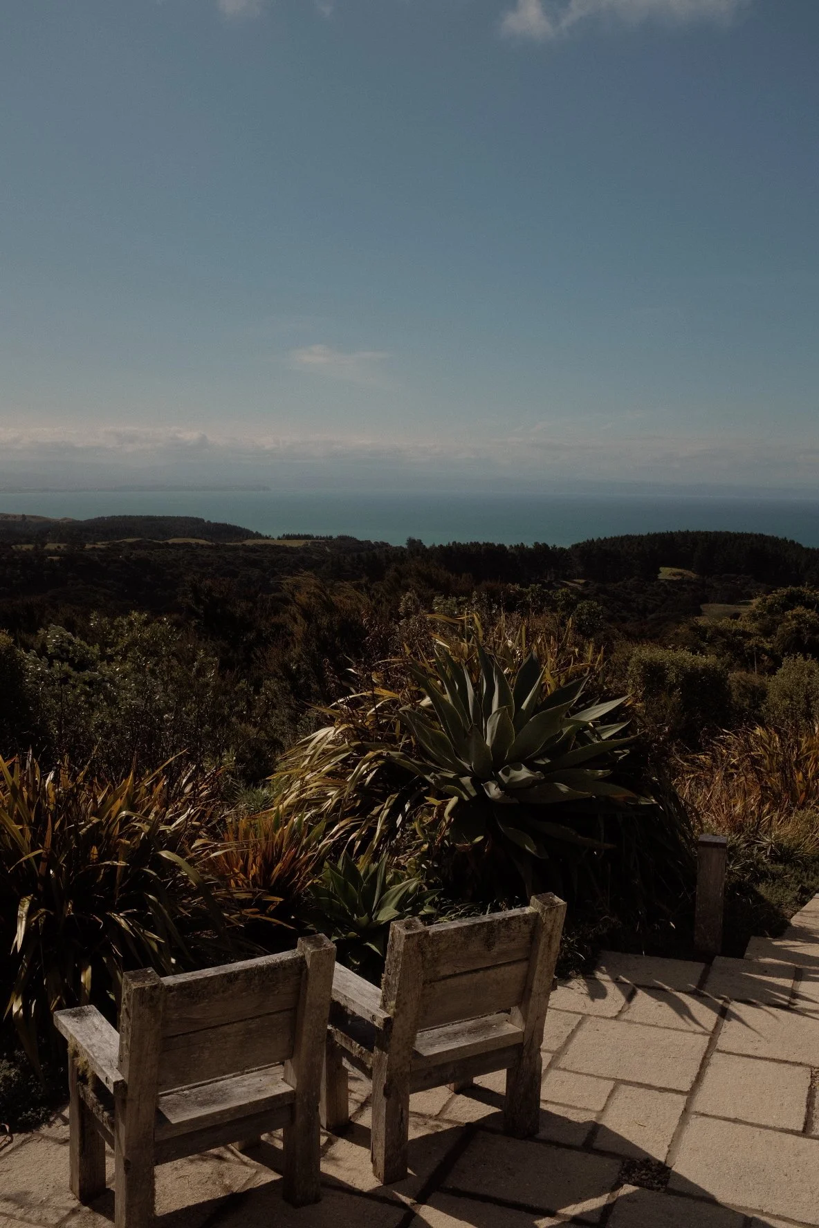Two weathered wooden chairs on a patio overlooking lush greenery, with a coastline visible in the distance under a clear sky.
