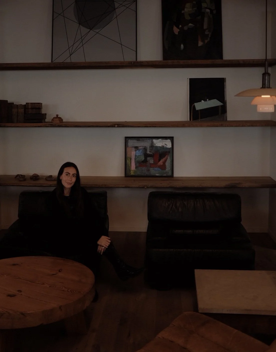 A woman sitting on a black leather sofa in a dimly lit room with wooden furniture and modern abstract art on the wall.