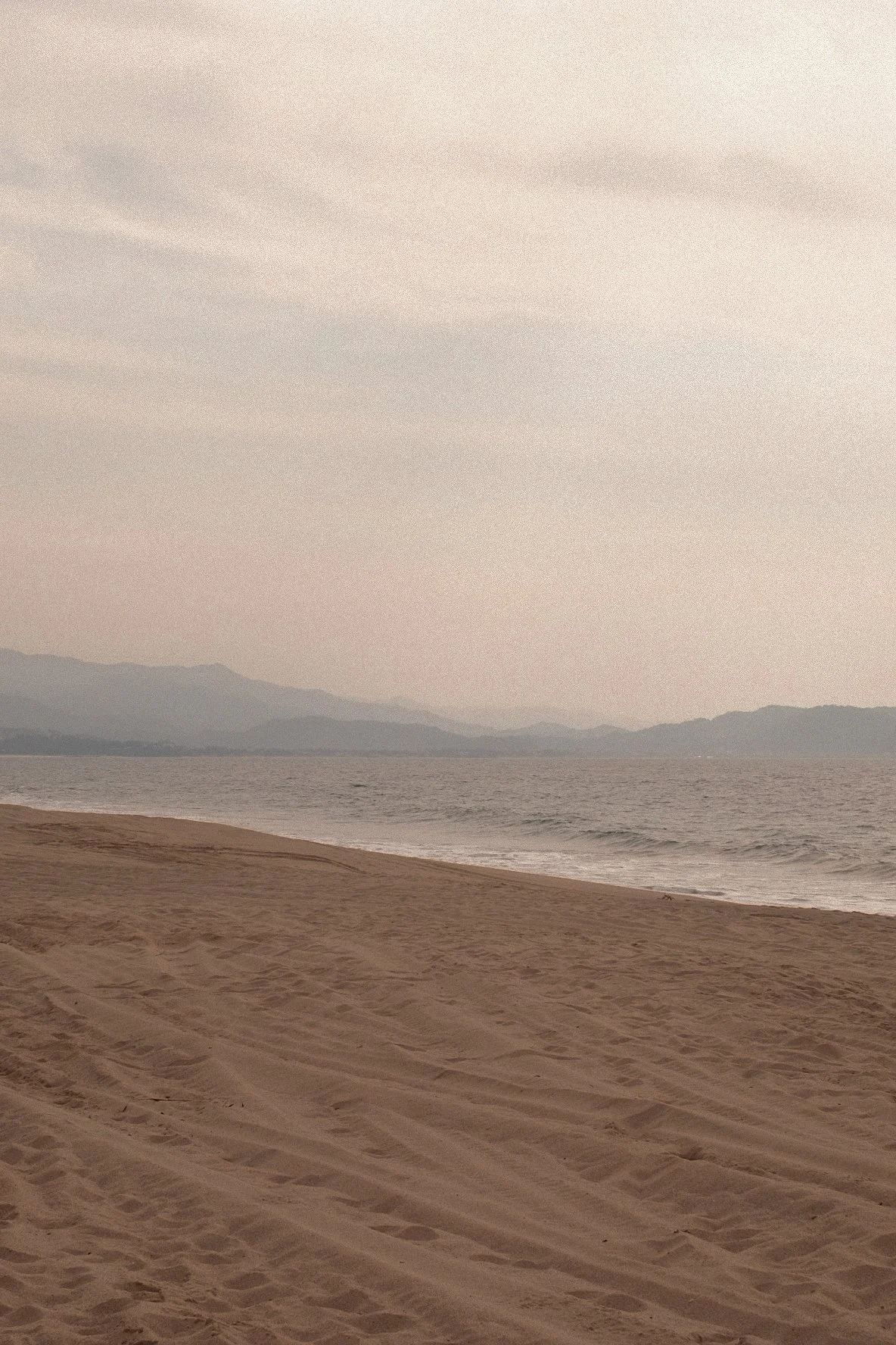 A sandy beach with tire tracks in the foreground, calm ocean waves in the middle, and distant mountains under a cloudy sky in the background.