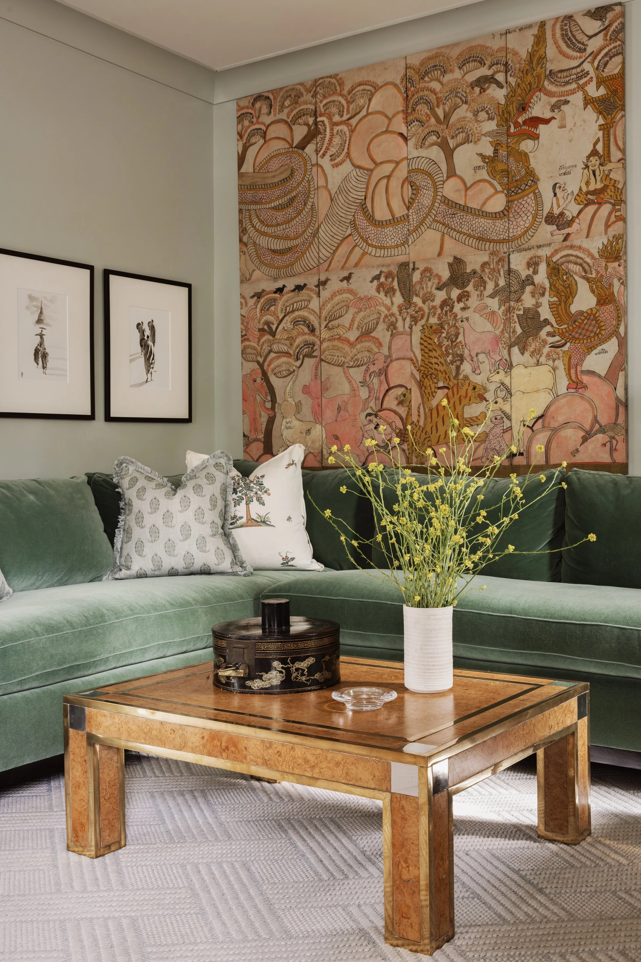 A living room with a green velvet sofa, decorative pillows, a wooden coffee table, a vase with yellow flowers, a large wall hanging with traditional Asian artwork, and two framed black and white photographs on the wall.