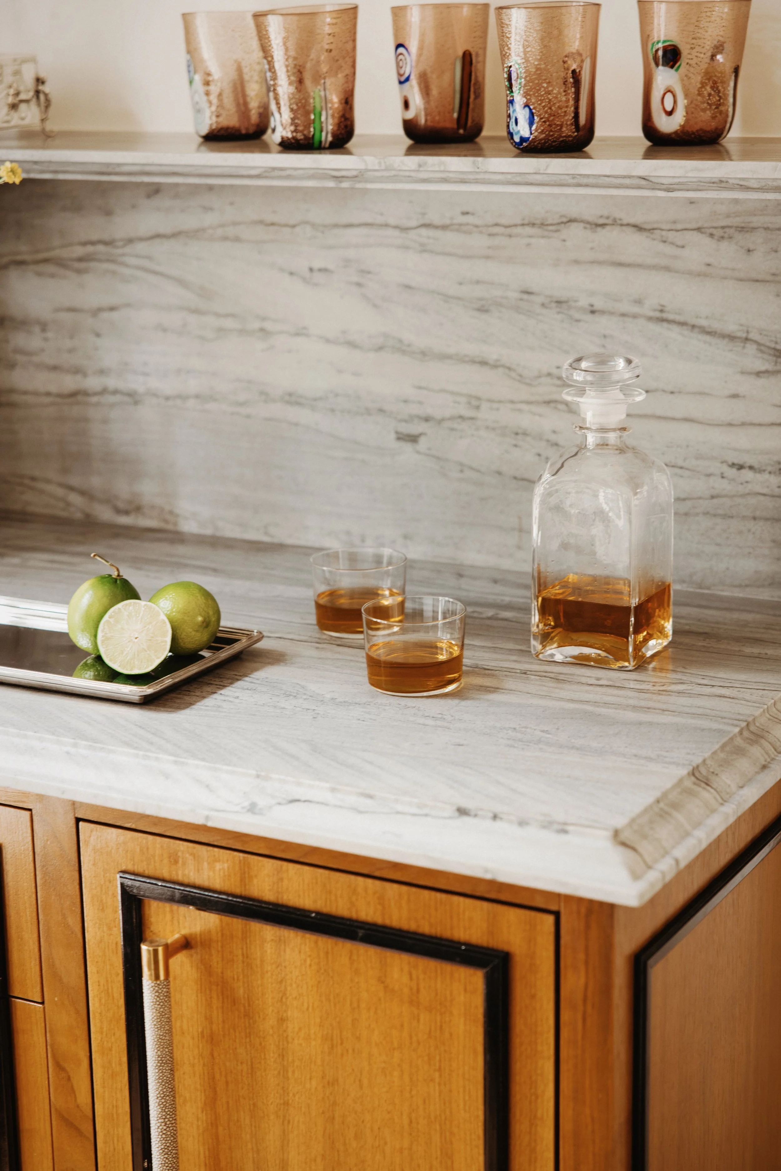 A kitchen countertop with a bottle of liquor, two glasses filled with a brownish drink, and a tray with two green limes and a sliced lime. The countertop is made of white marble, and the cabinets below are wooden with black accents.