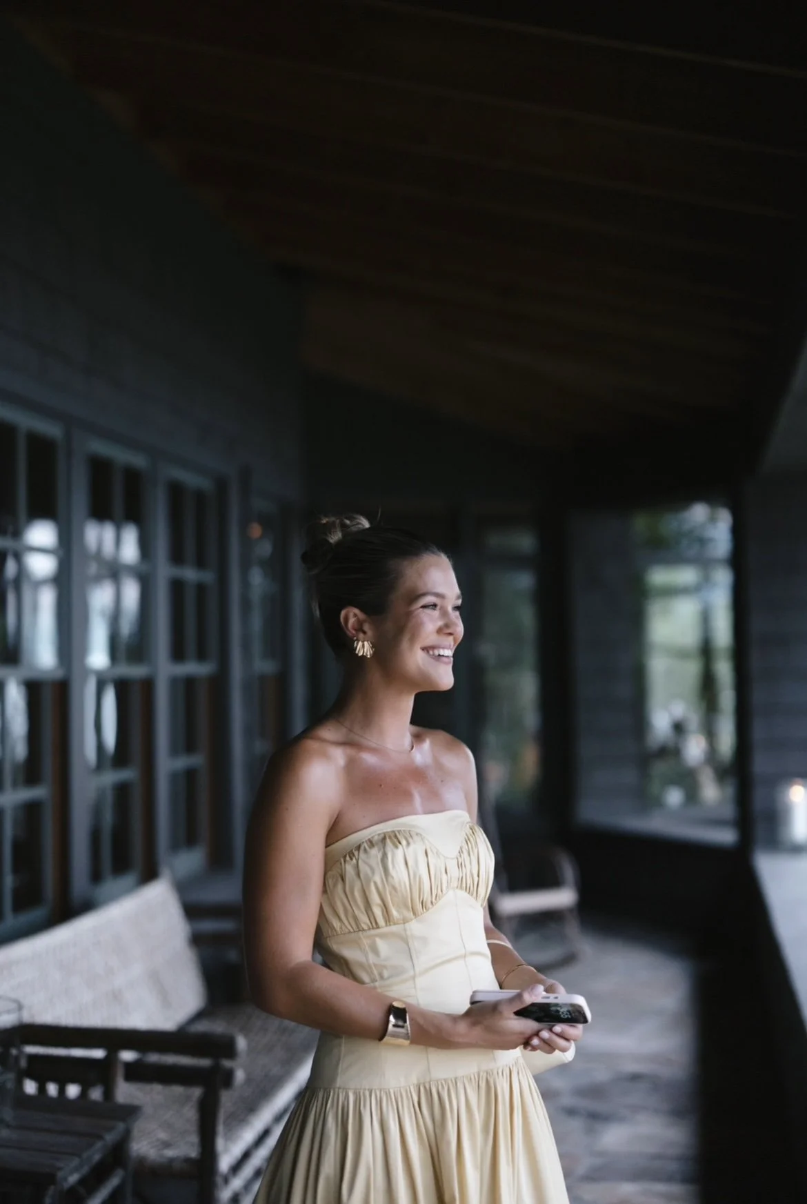 Woman in a strapless yellow dress smiling indoors, holding a smartphone, with a dimly lit background of windows and seating.