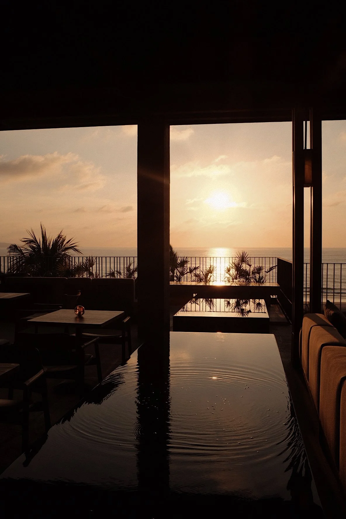 Sunset view over the ocean seen from a covered patio area with outdoor furniture, plants, and railing.
