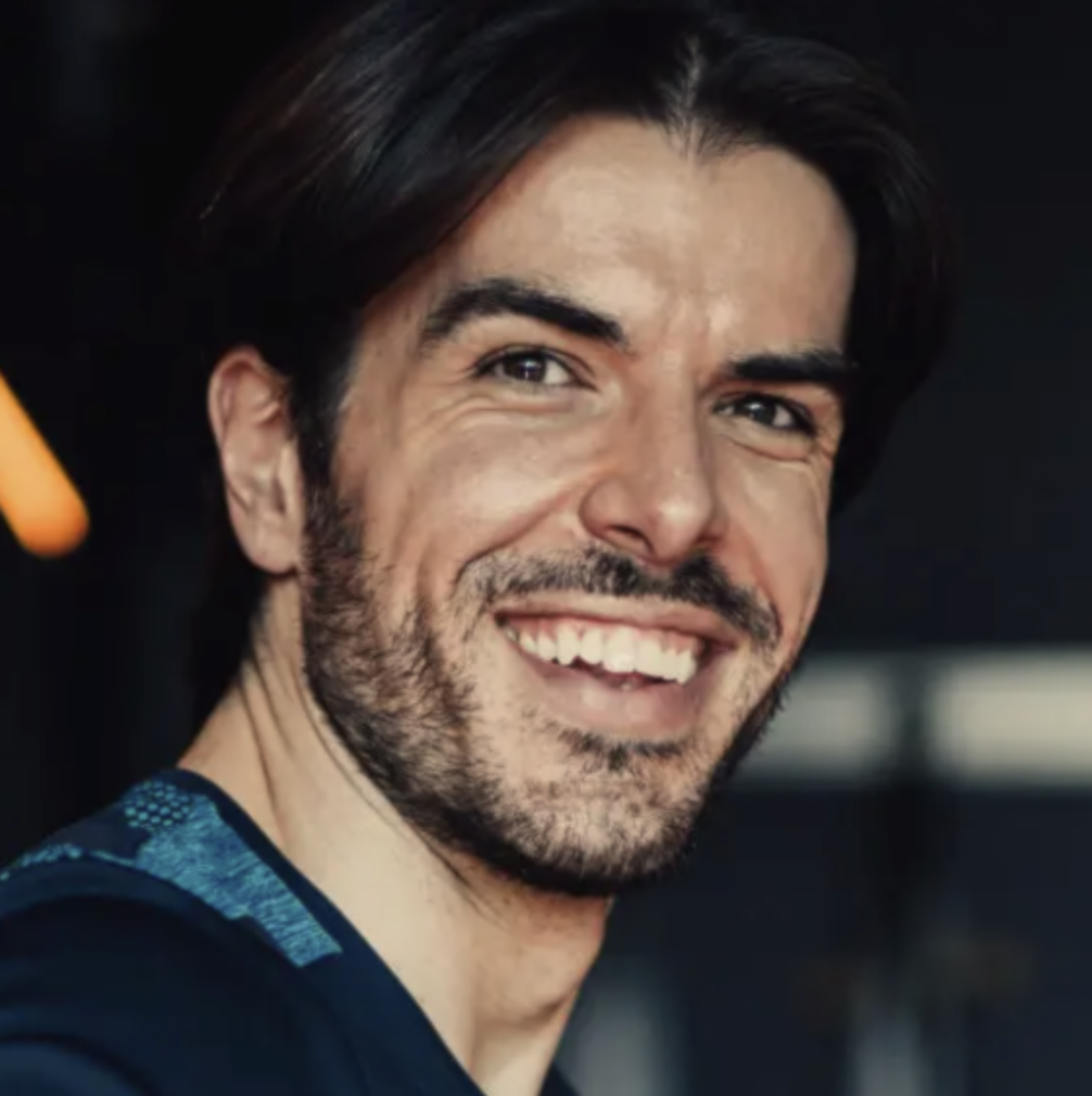 A man with dark hair and a beard smiling at the camera, wearing a dark shirt, with a blurred background.