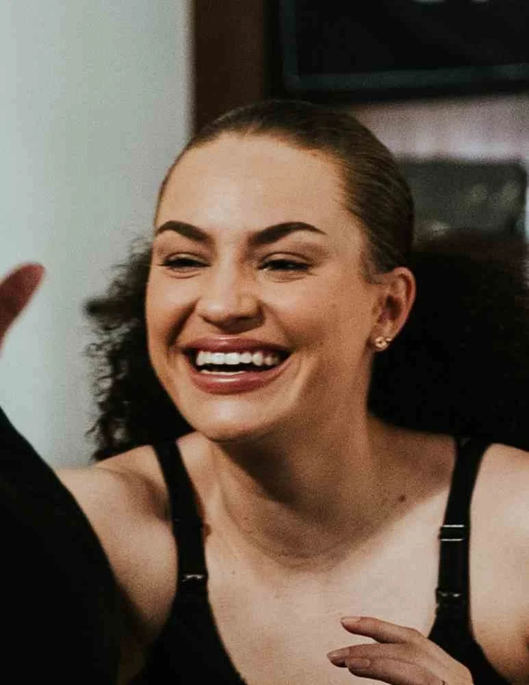 A woman with dark curly hair smiling and waving, wearing a black top with thin straps.