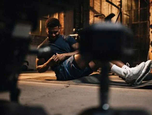 A man is sitting on the floor of a gym, smiling, surrounded by gym equipment, and appears to be relaxing after a workout.