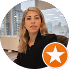A woman with blonde hair wearing a black blazer, sitting indoors with large windows showing buildings outside.