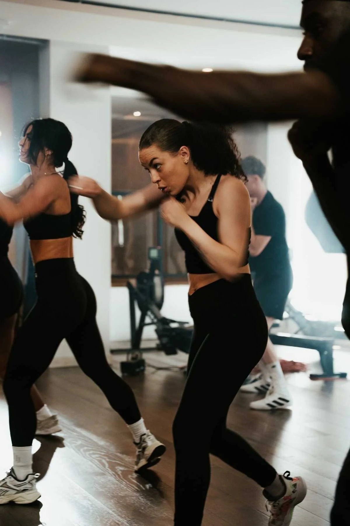Women participating in a fitness class, engaging in a synchronized workout or dance, wearing athletic wear, in a studio with wooden floors and mirrors.