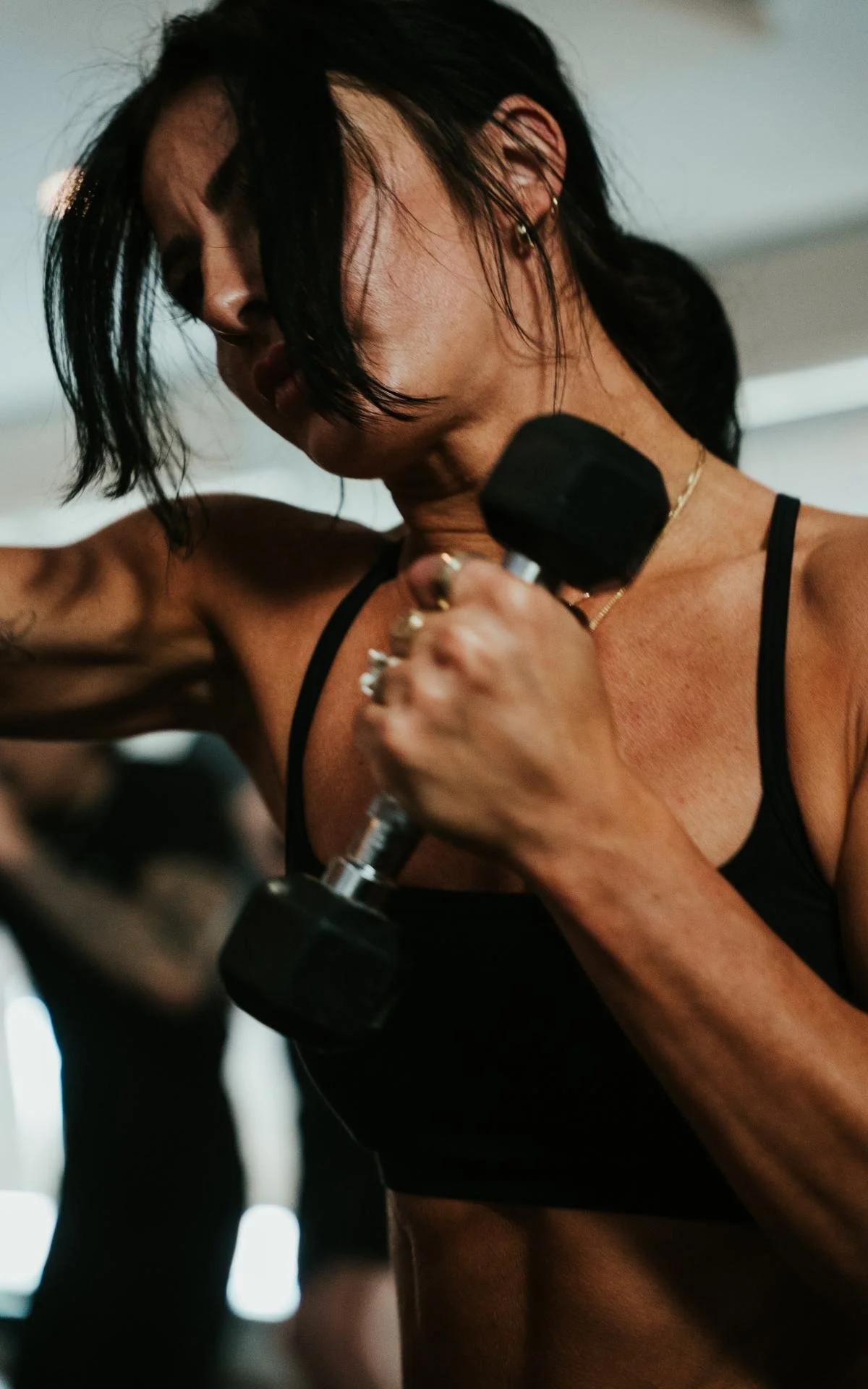 A woman with dark hair and earrings in a black sports bra, lifting a black dumbbell during a workout.