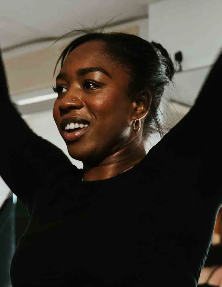 A woman with dark skin and black hair tied back, wearing a black top, smiling and raising her arms.