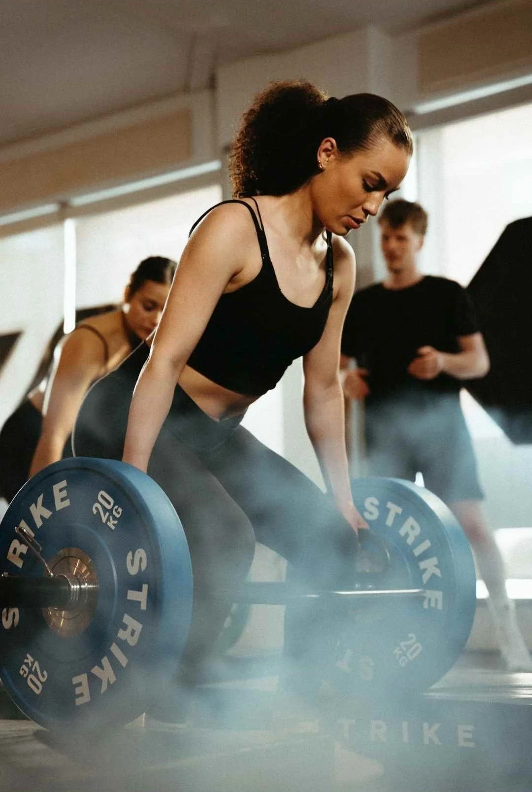 A woman with curly hair in a ponytail wearing a black sports bra and leggings lifting a 20 kg blue barbell in a gym, with two other people in the background.